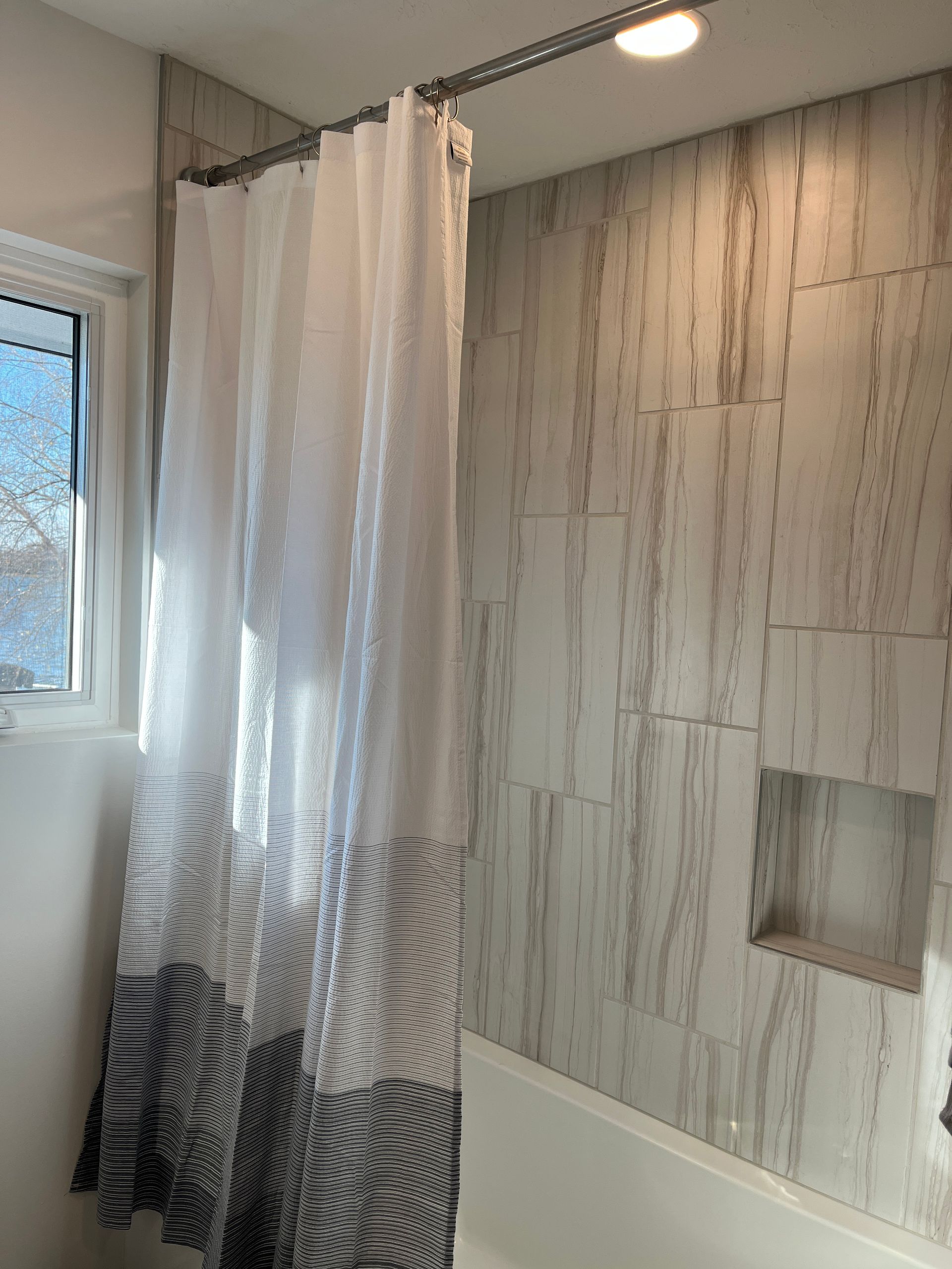 A bathroom with a white shower curtain, gray and white tile, and a small window. There is a recessed shelf in the wall.