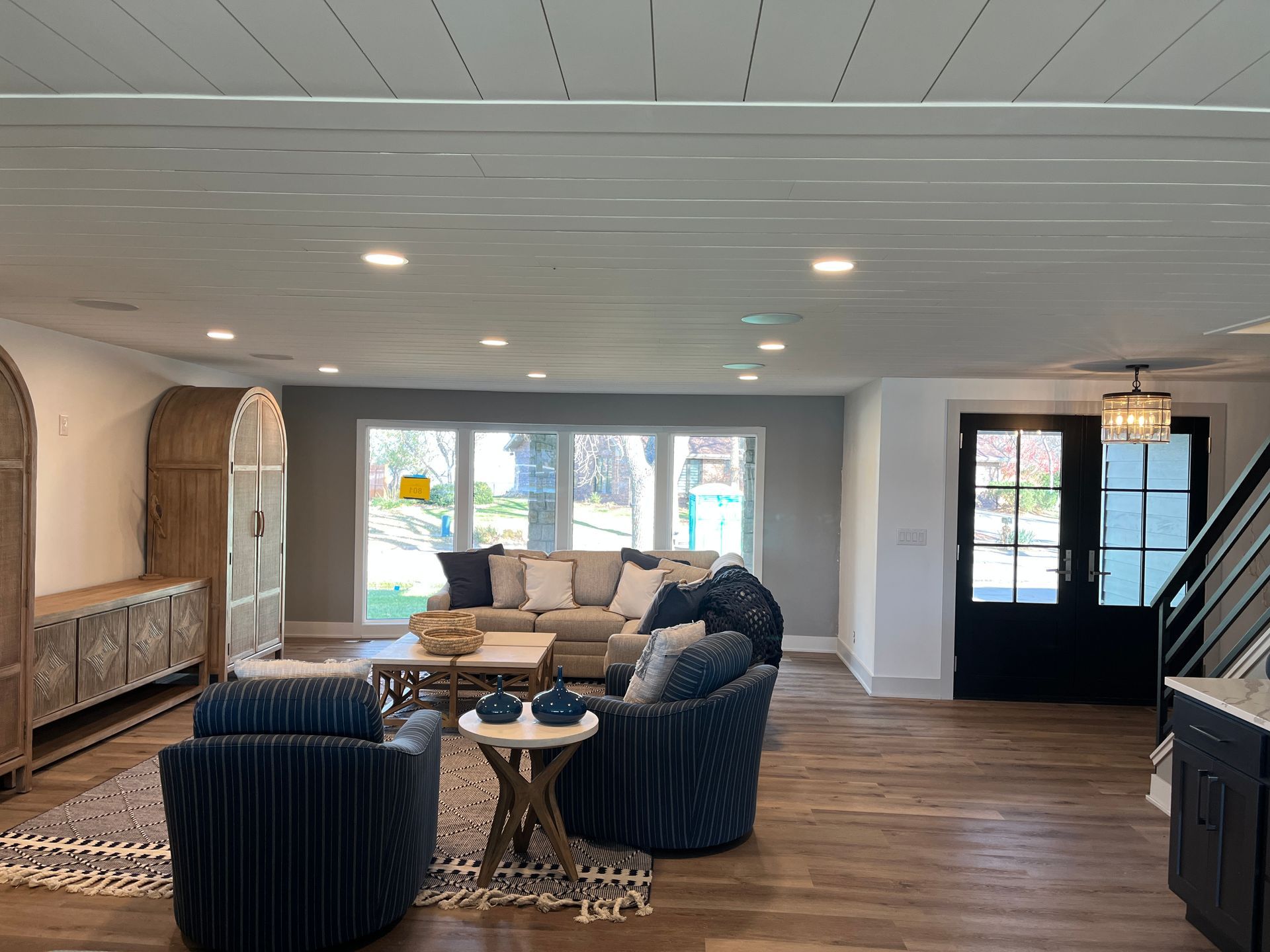 A living room with blue armchairs, a beige couch, and a white-paneled ceiling. Large windows overlook a backyard.