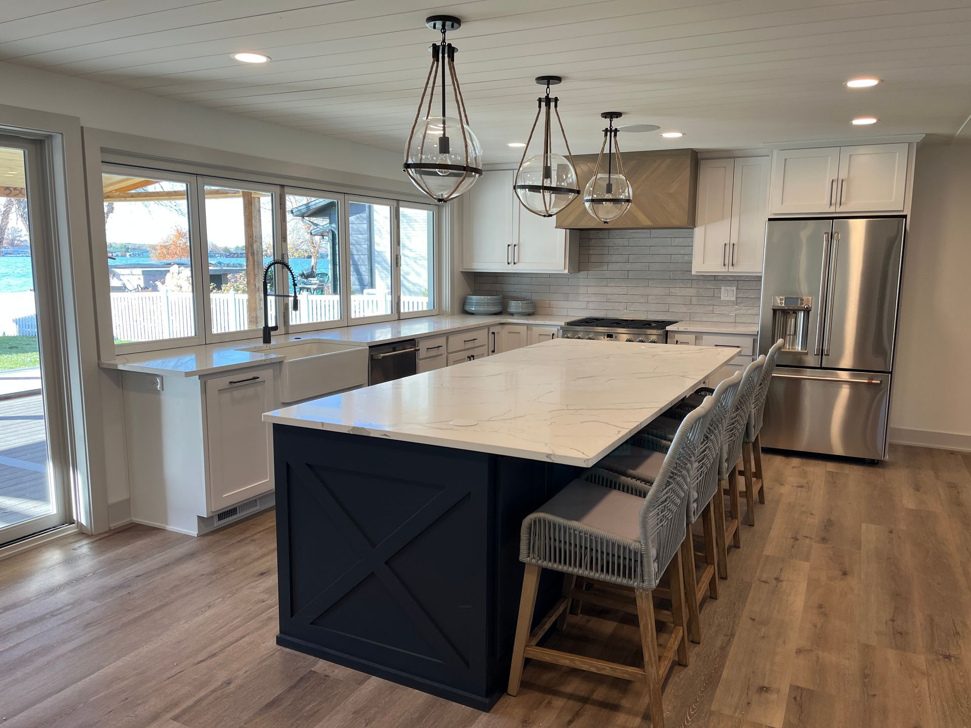 Modern kitchen with a navy island, white countertops, and stainless steel appliances. Windows overlook a waterfront view.