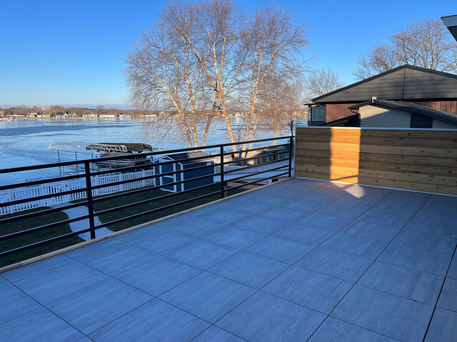 A rooftop deck overlooking a frozen lake. The deck is gray, with black railing, and a wooden privacy fence.