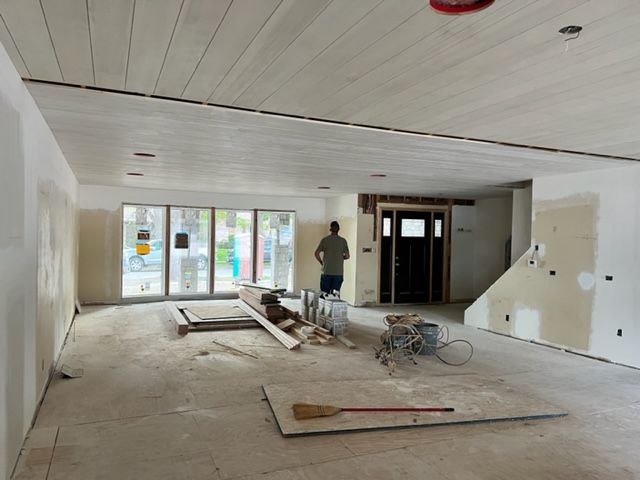 Interior of a room under construction: white walls, wood plank ceiling, large windows, and a man standing in the middle.