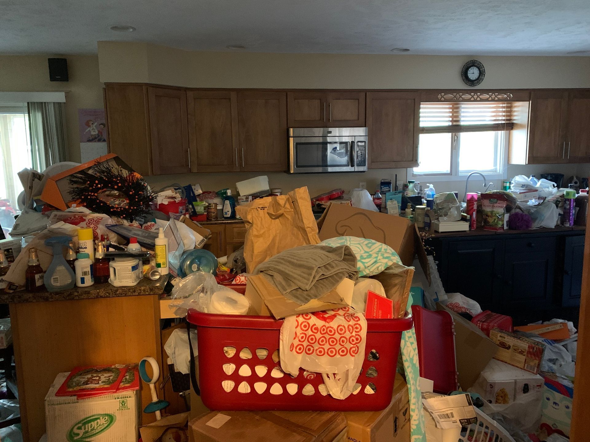 A cluttered kitchen with piles of items covering countertops, a red basket in the foreground, and brown cabinets.