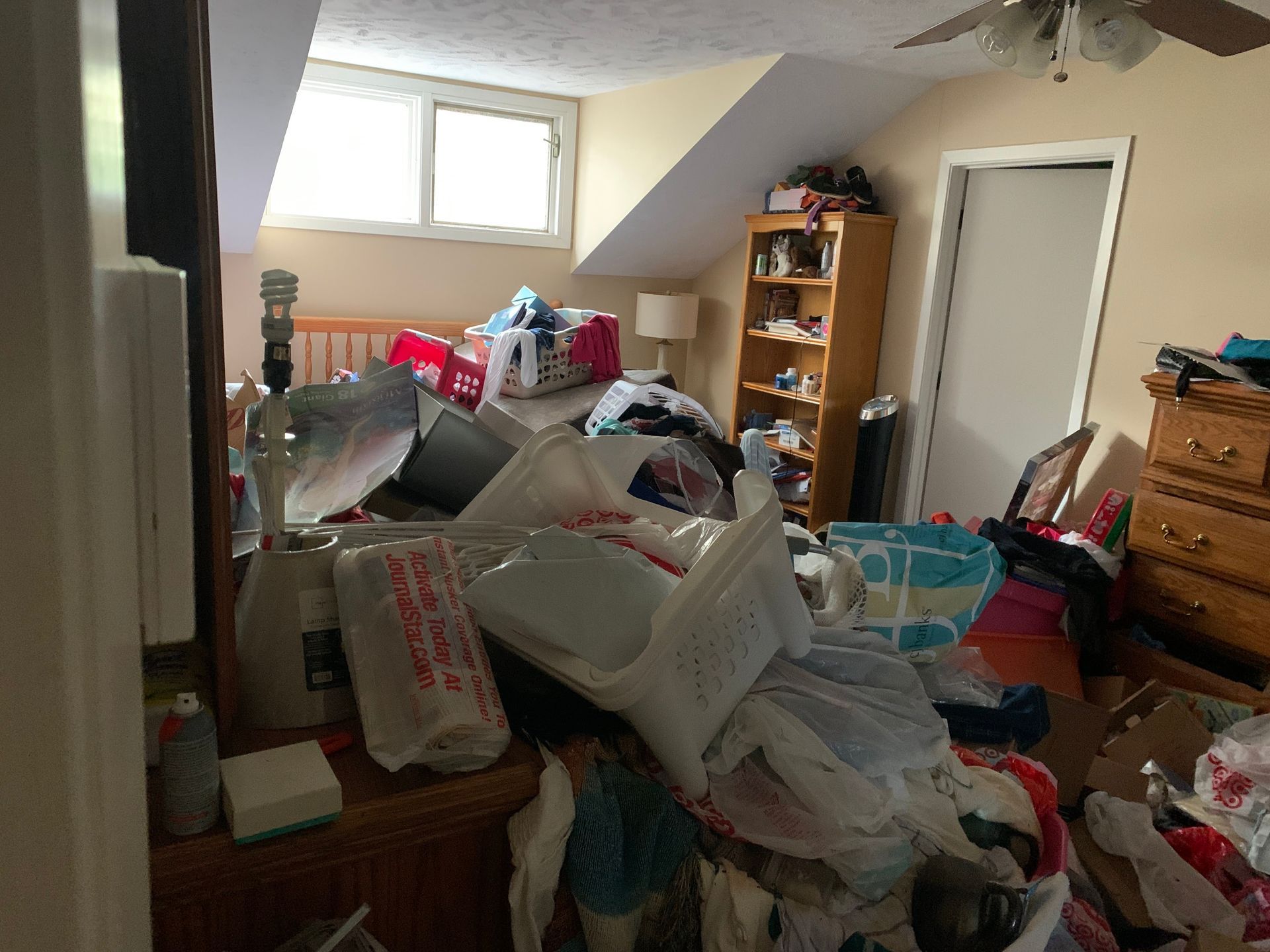 A cluttered bedroom with clothing, bags, and items piled on the bed, floor, and furniture. A window and door are visible.