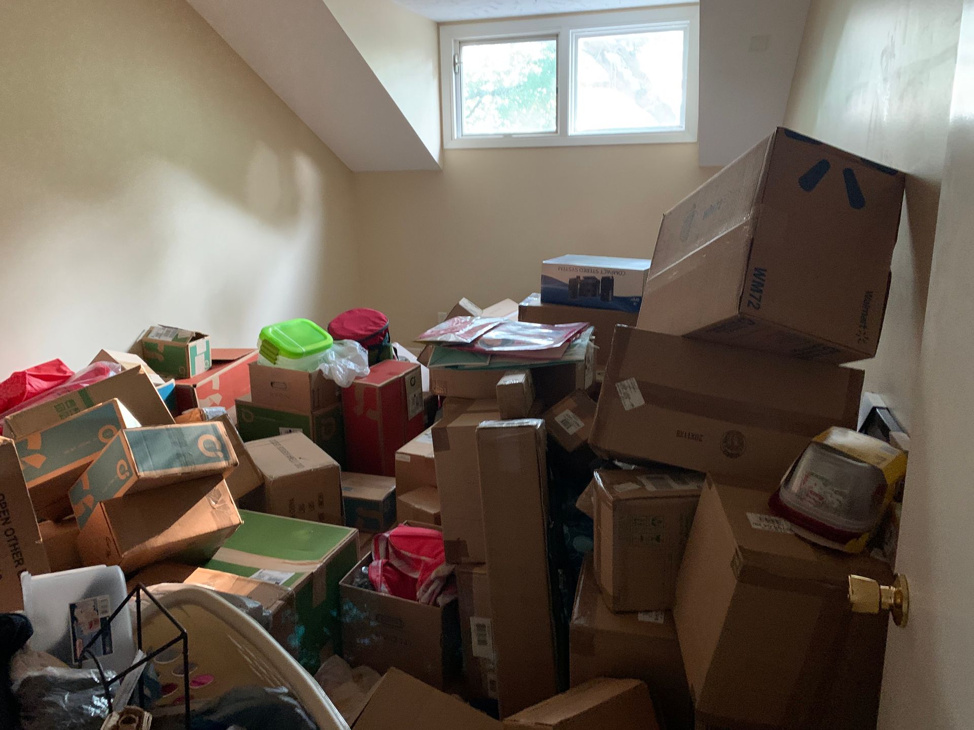 Pile of cardboard boxes and miscellaneous items filling a room with a small window, indicating a cluttered storage space.