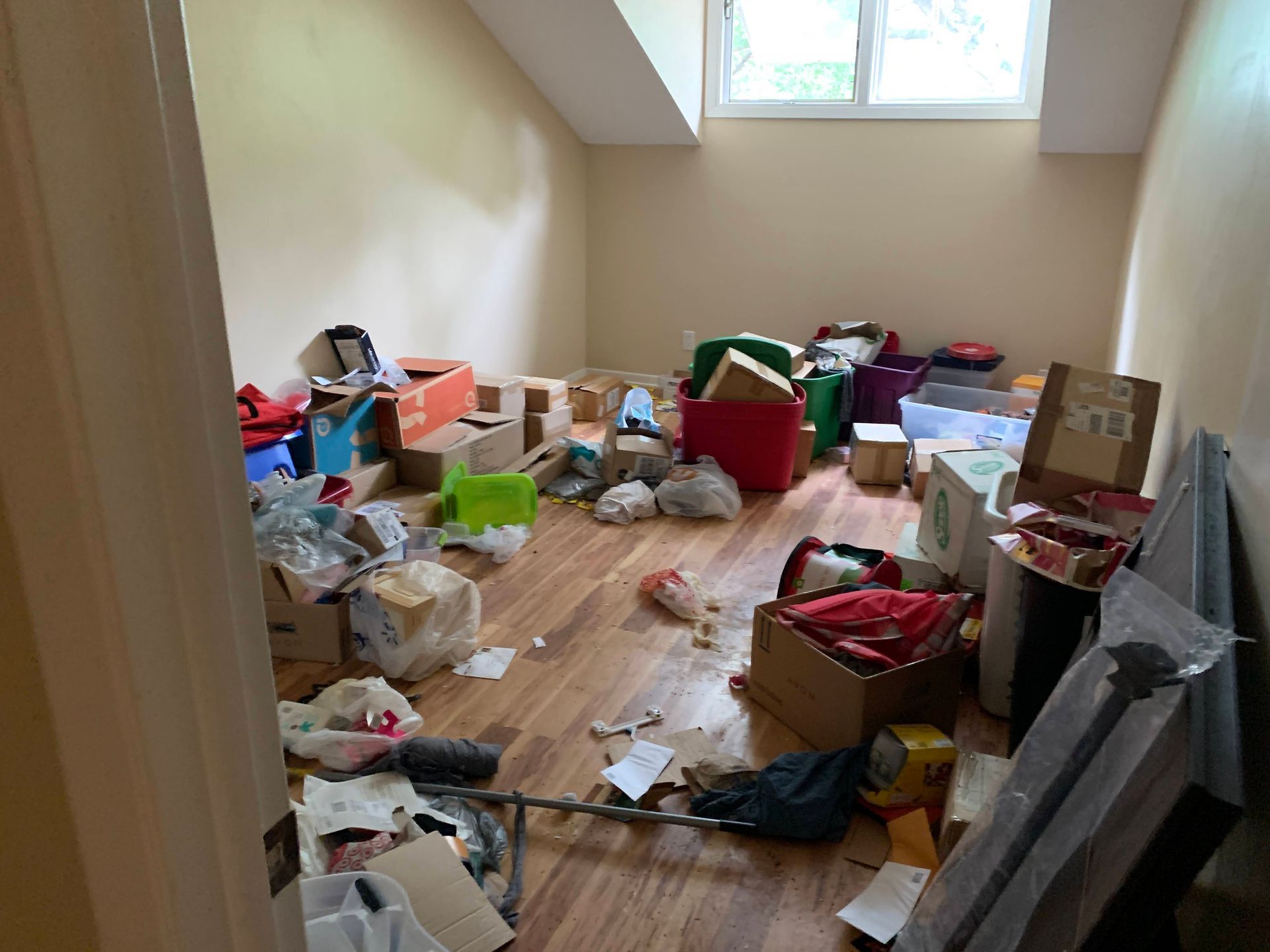 A cluttered room with many boxes, bags, and debris on the floor. Light-colored walls and wooden flooring.