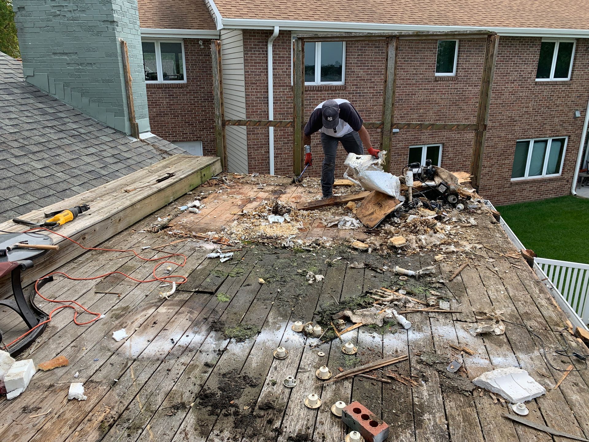 Person removing debris from a damaged wooden deck. In the background, a multi-story brick building with windows.