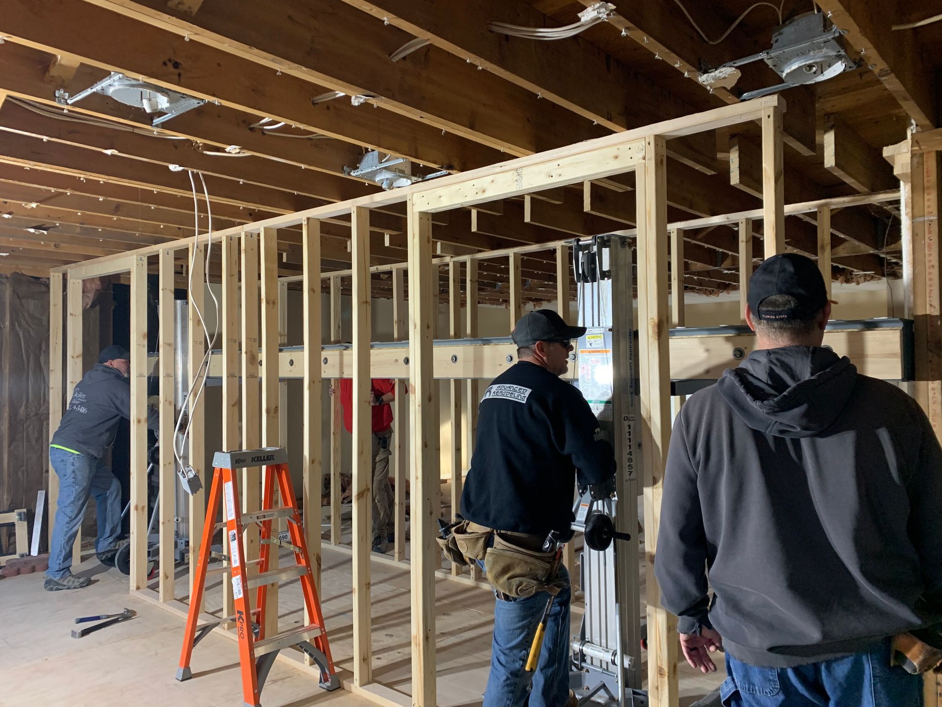 Construction workers framing a wall indoors. Three men are working, using tools and a ladder. 