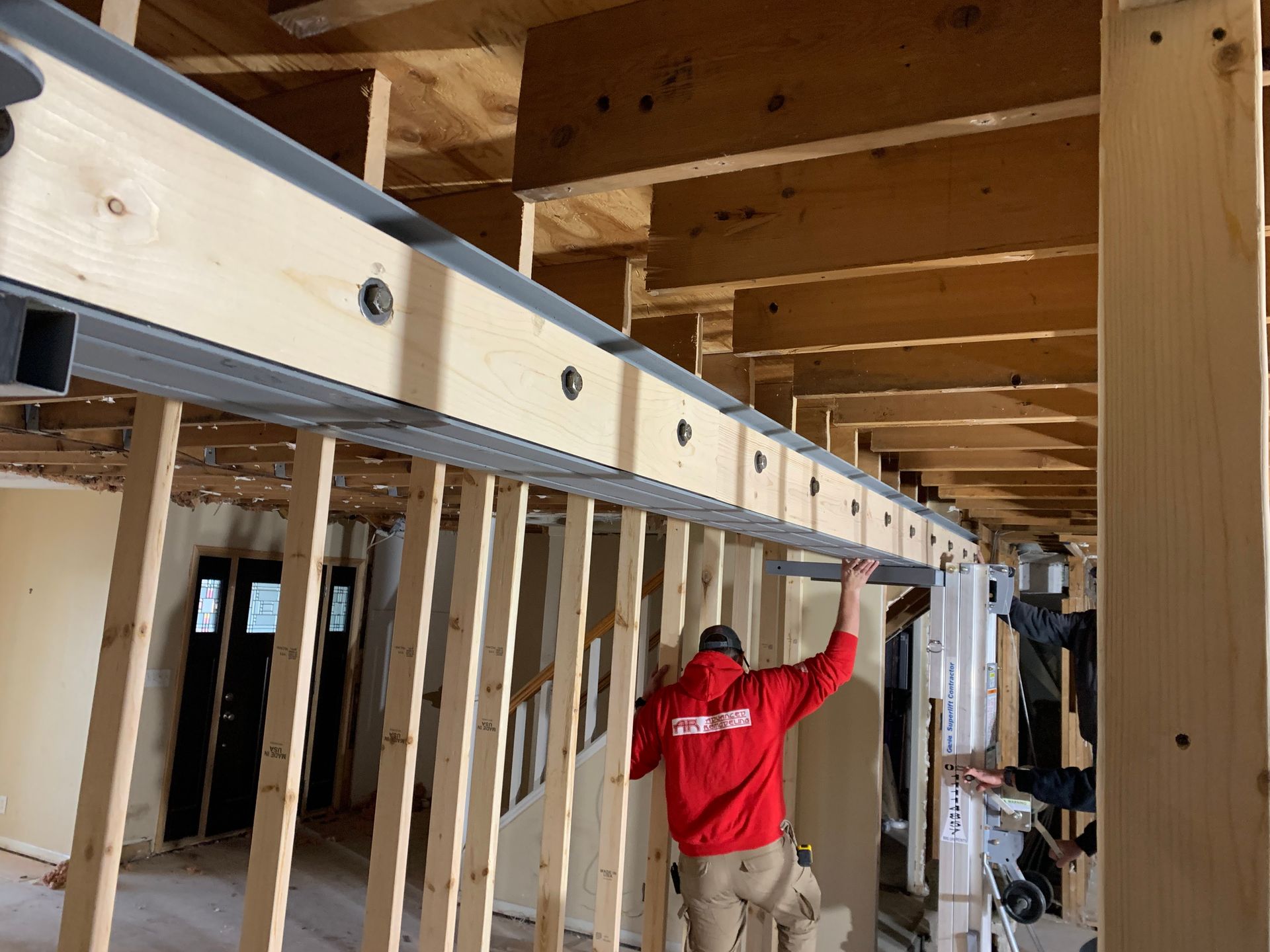 A person in a red jacket works on a construction project, possibly installing a metal beam, with wood framing visible.
