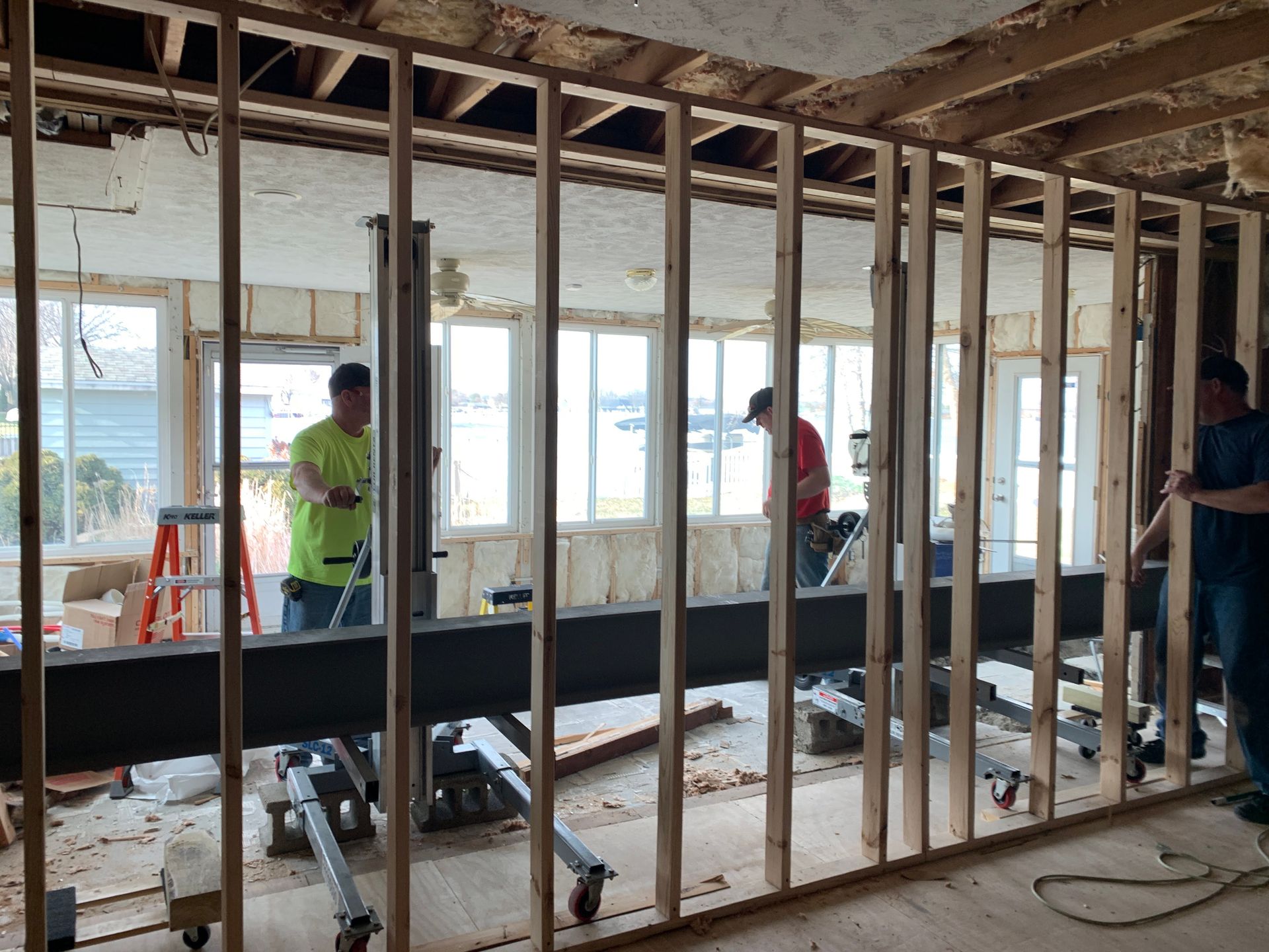 Construction workers installing a steel beam. Wooden framing is in place with windows in the background.
