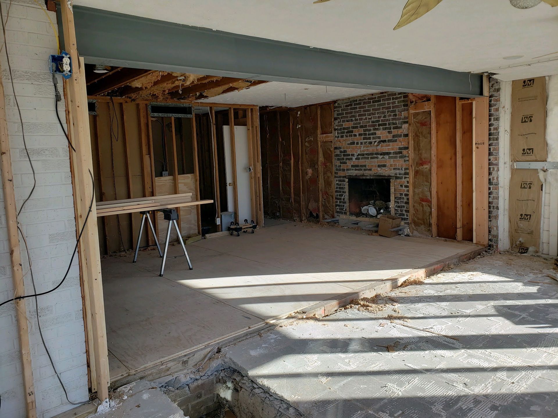 Interior of a house under renovation with exposed studs, a steel beam, and a brick fireplace. Sunlight streams in.