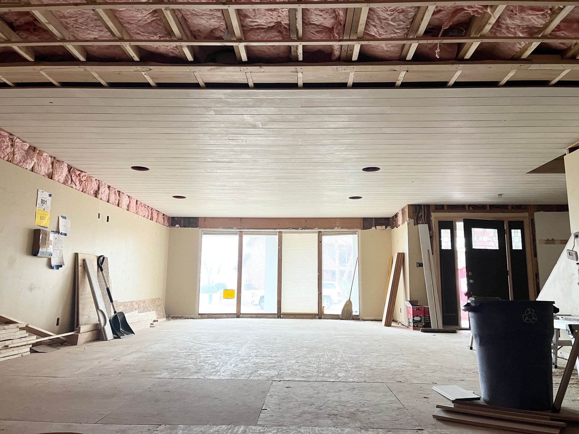 Interior of a room under renovation, showing exposed ceiling beams, insulation, and open doorway/sliding glass doors.