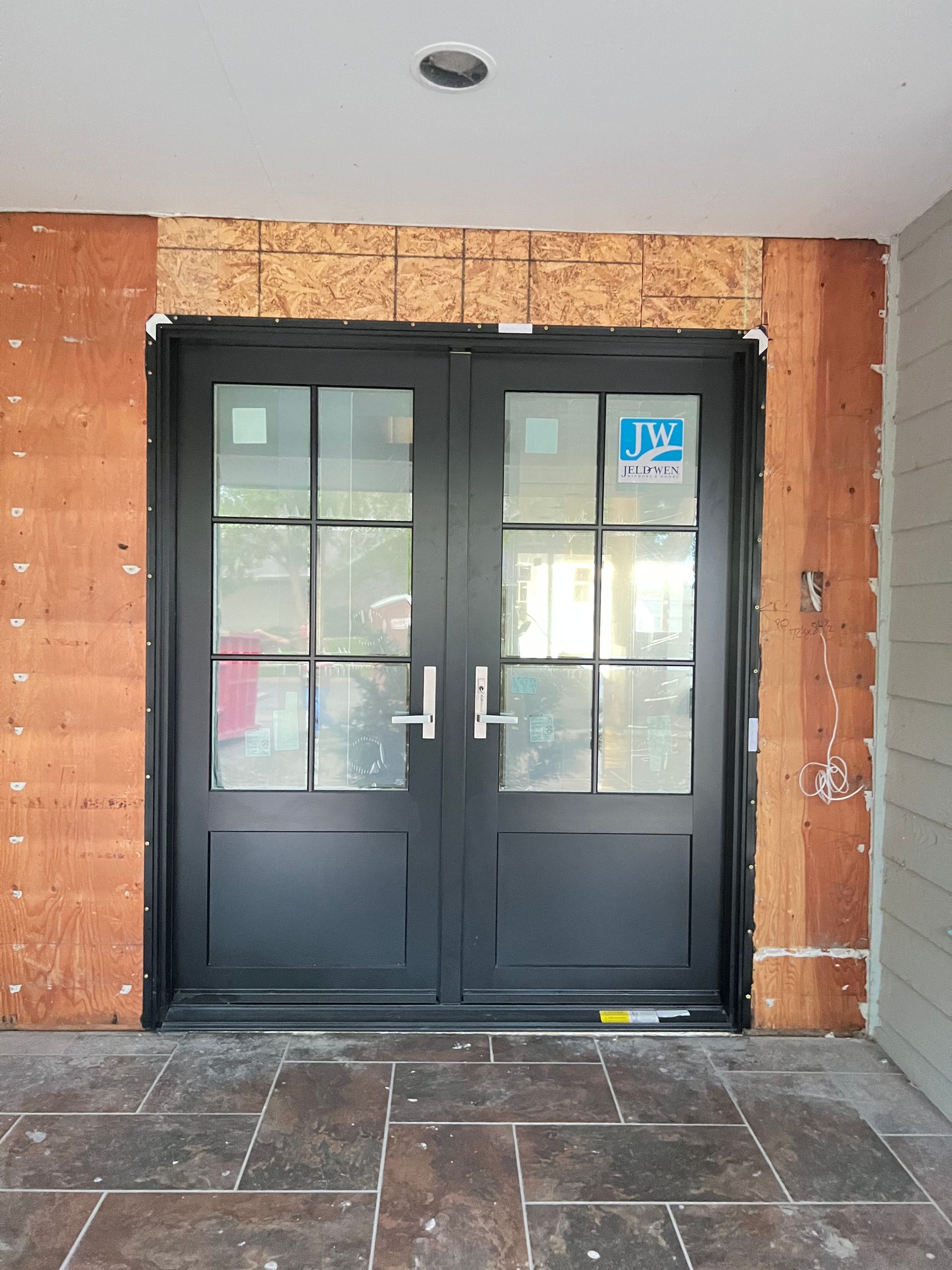 Black double doors with glass panes, set in a doorway with unfinished brown framing. Paved floor.