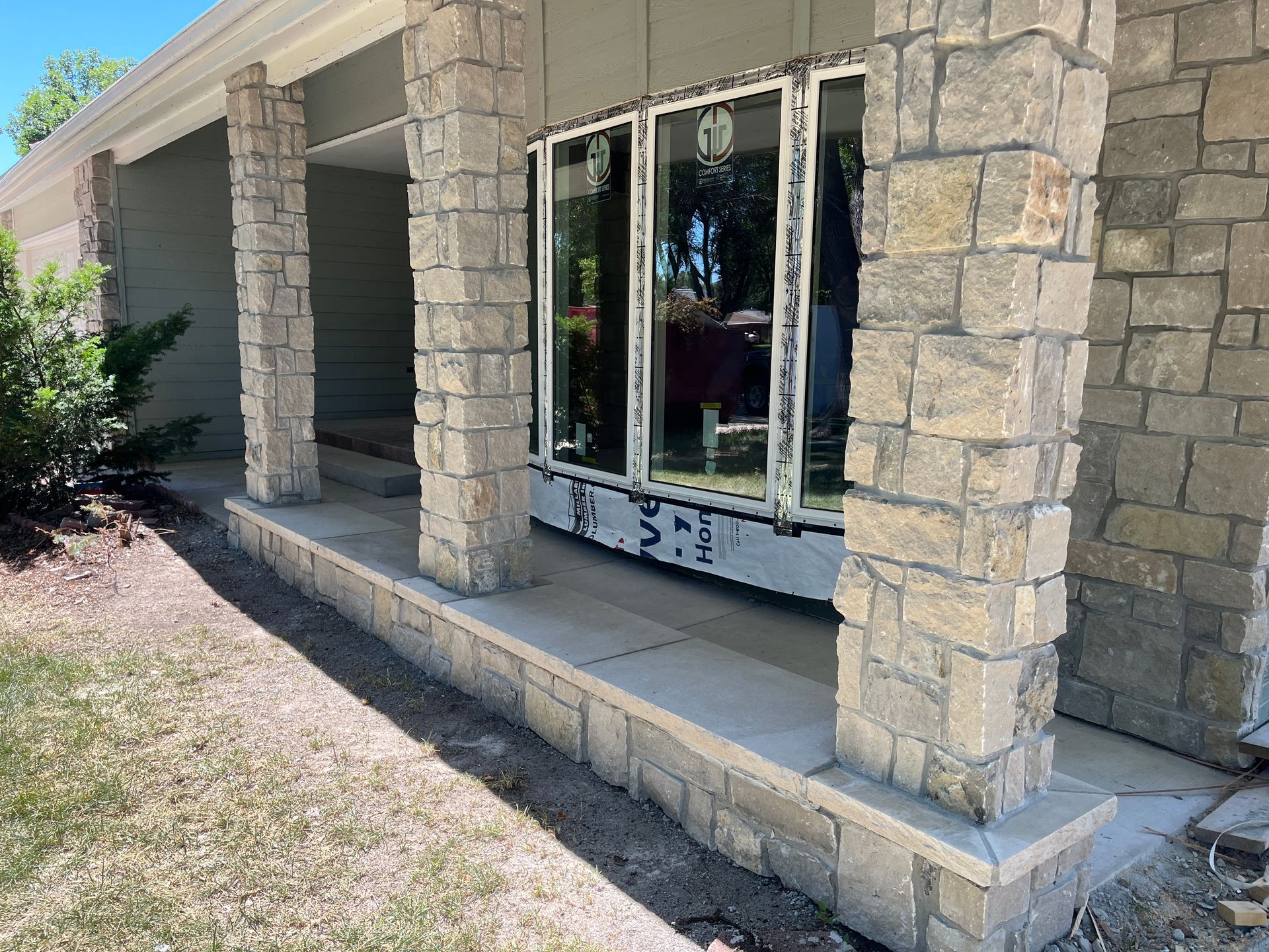 Stone-faced pillars frame a porch with a gray wall and a window. The porch is bordered by a low stone wall.