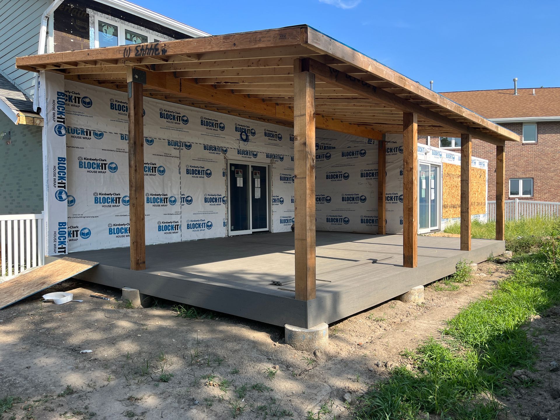 Construction of a covered patio attached to a house. Concrete floor, wood supports and roof, house wrap on exterior walls.