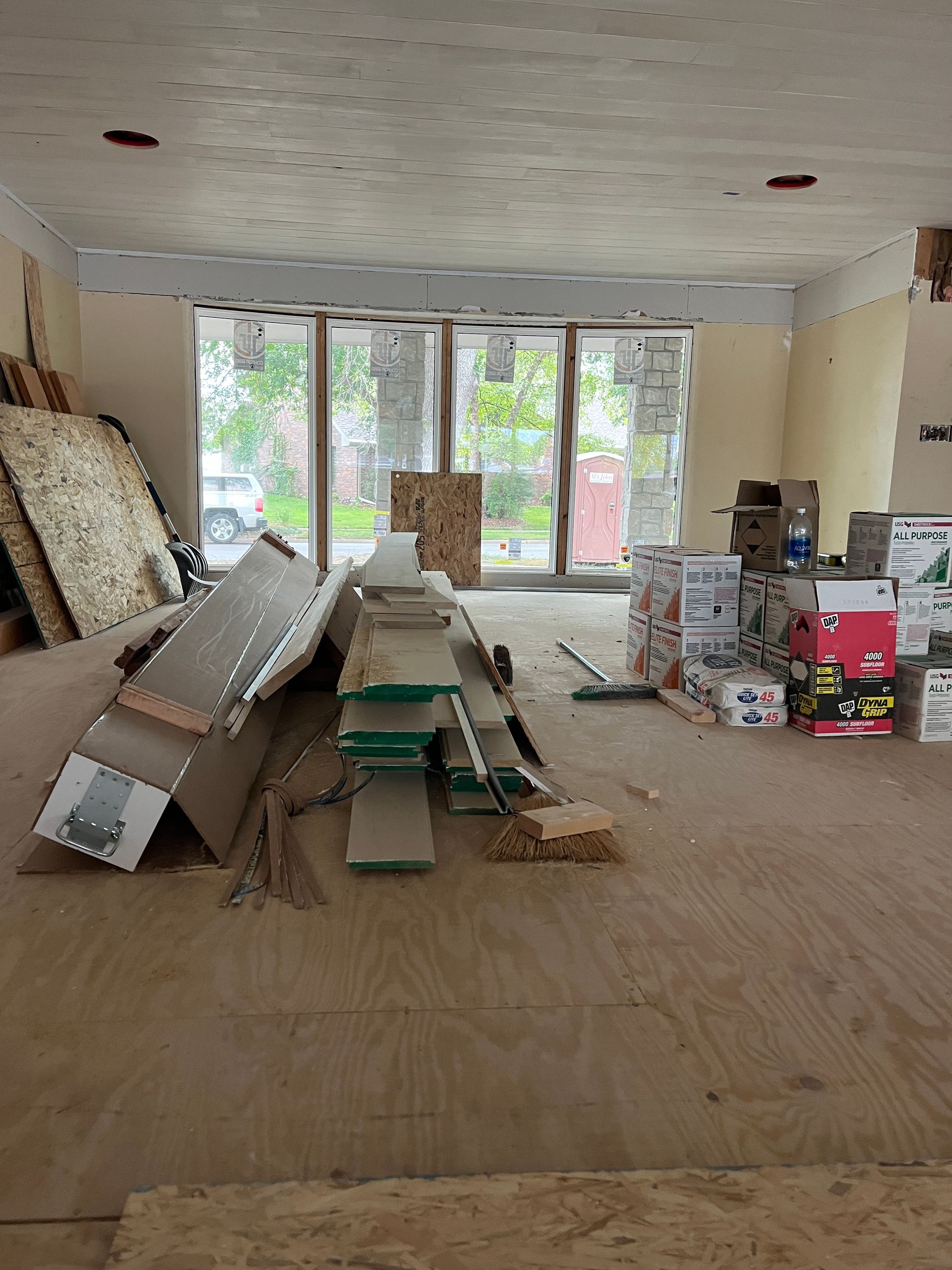 Interior of a room under renovation with plywood floors, construction materials scattered about, and a large window.