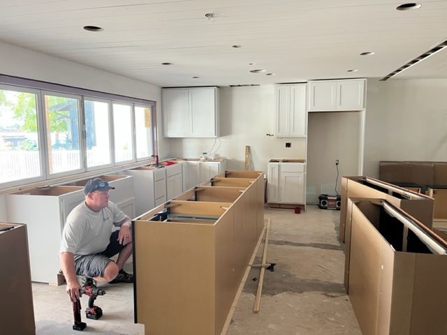 A person kneels while installing white kitchen cabinets. The room has bare walls, a large window, and unfinished flooring.