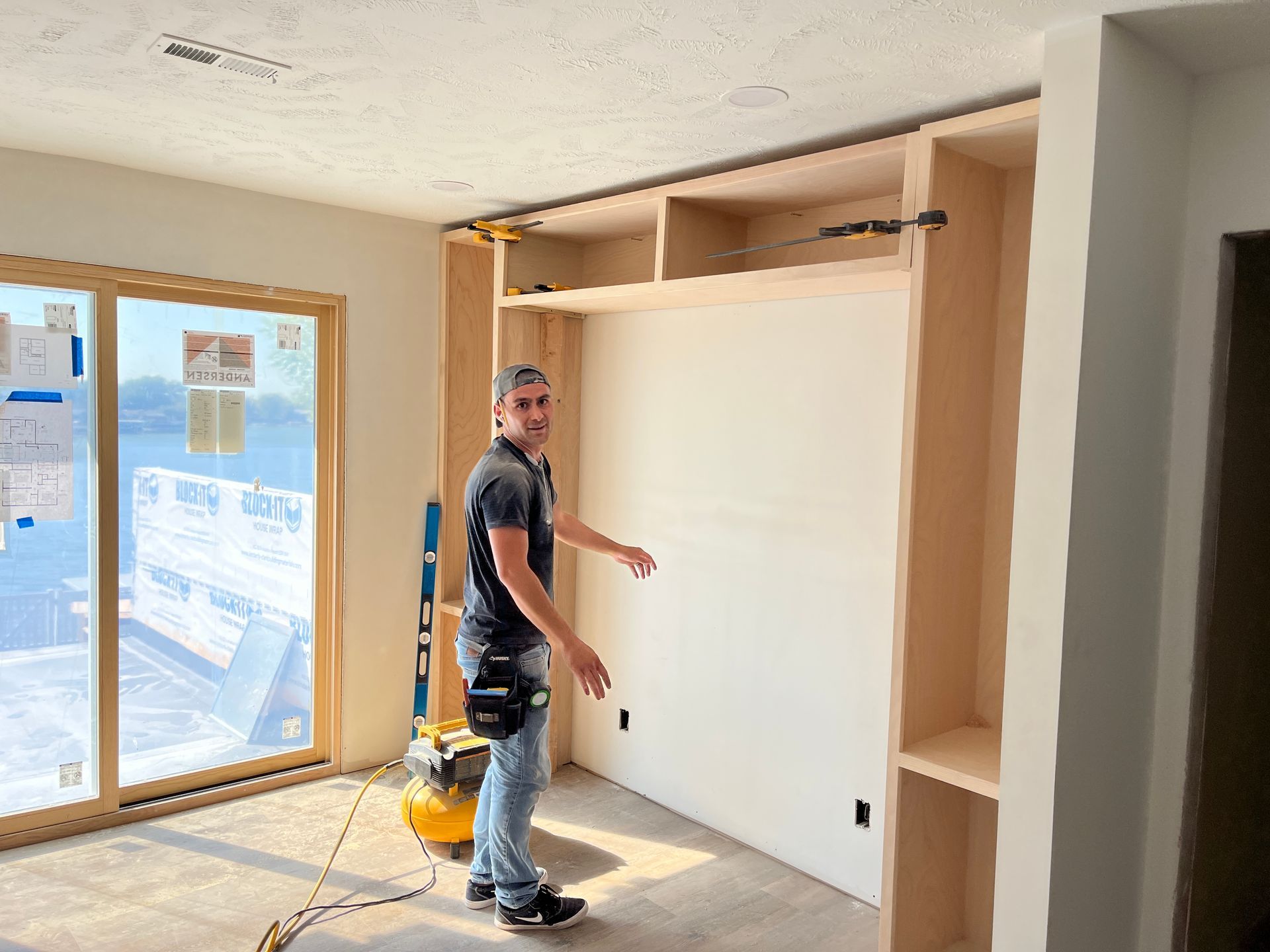A man installing built-in cabinetry in a room with large windows. He points towards a wall unit with open shelves, smiling.