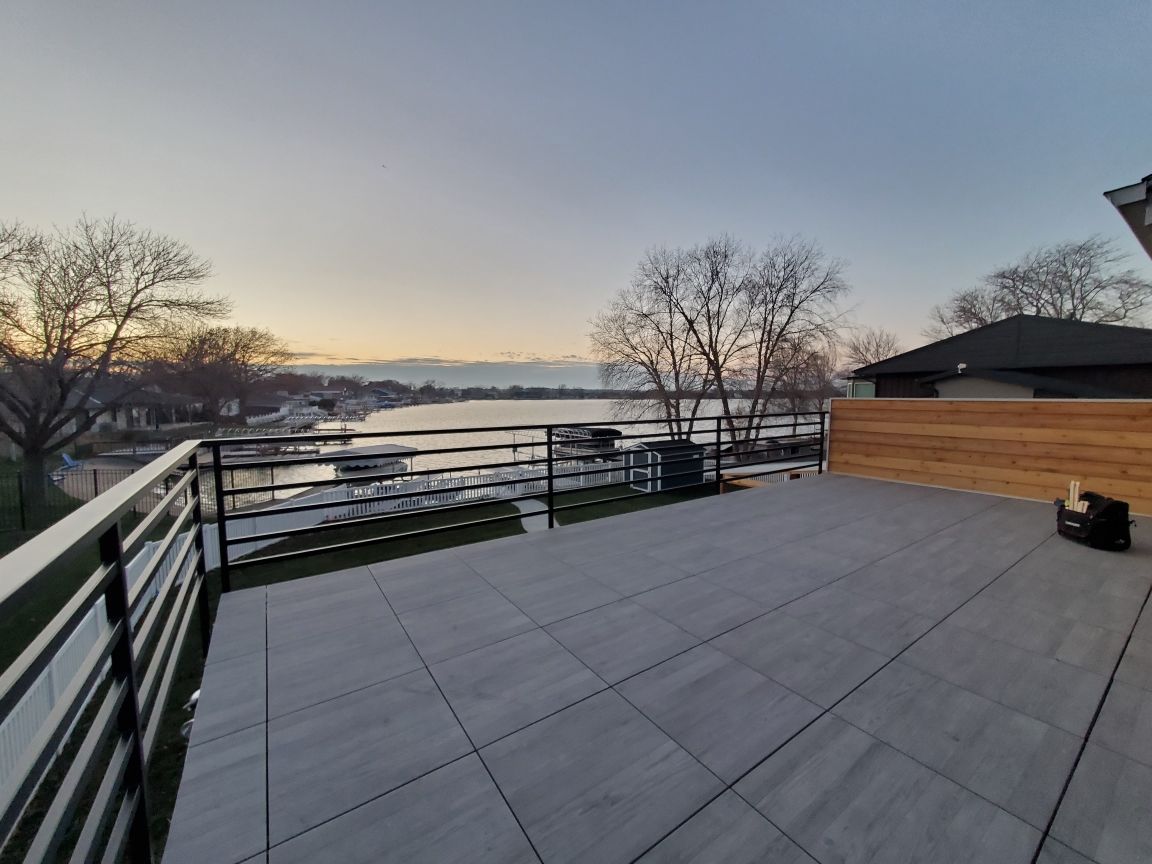 Large outdoor deck with gray tiled floor overlooking a lake at dusk. Black metal railing, wooden fence, are also visible.