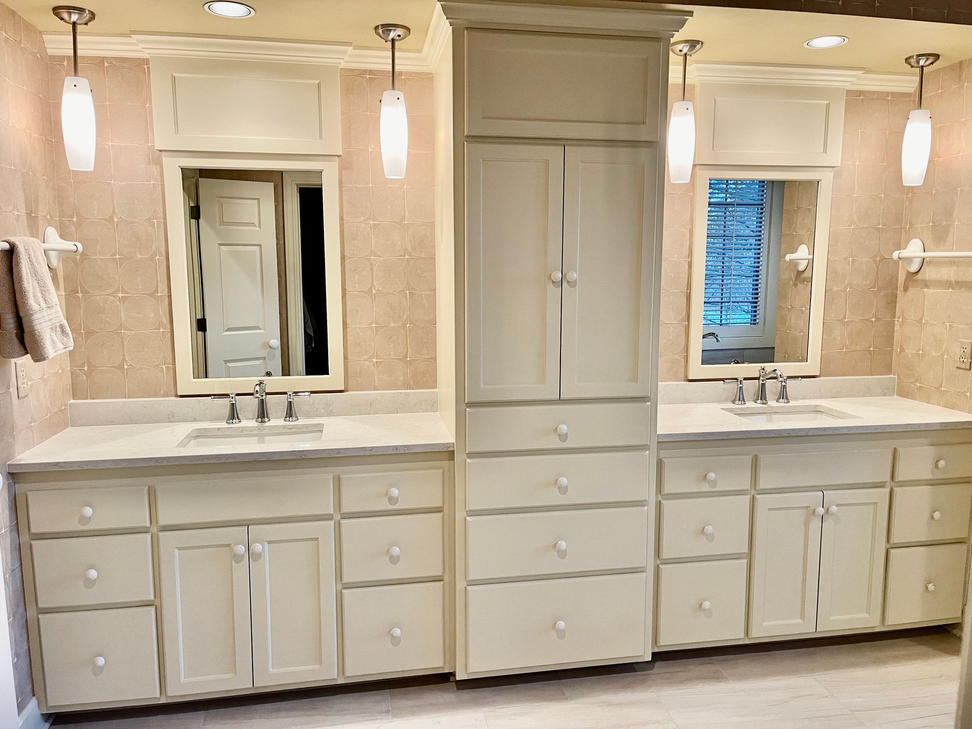 Two cream-colored bathroom vanities with mirrors and a tall storage cabinet. Neutral-toned walls and overhead lighting.