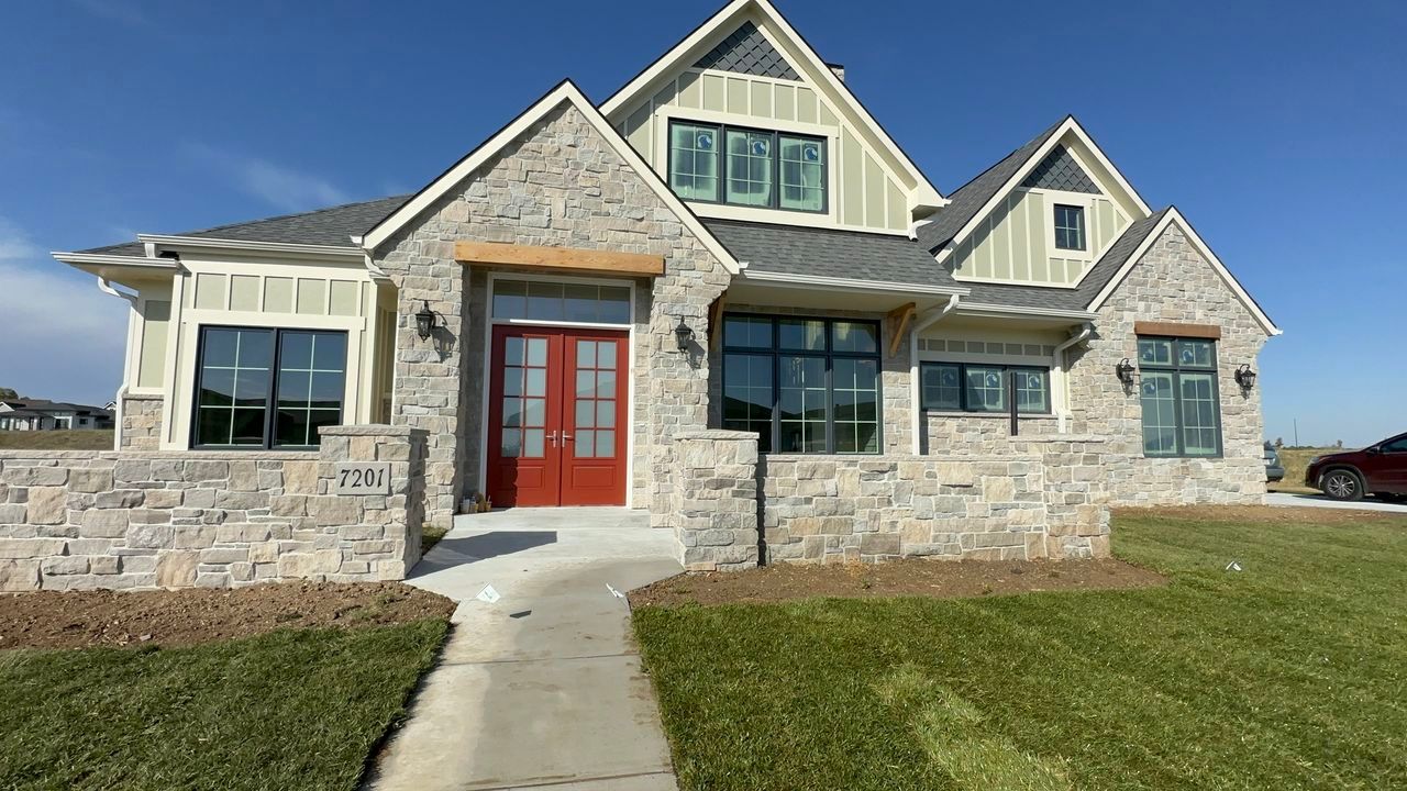 Stone-faced house with red door, black windows, and gray roof against a blue sky, with a concrete walkway and green lawn.