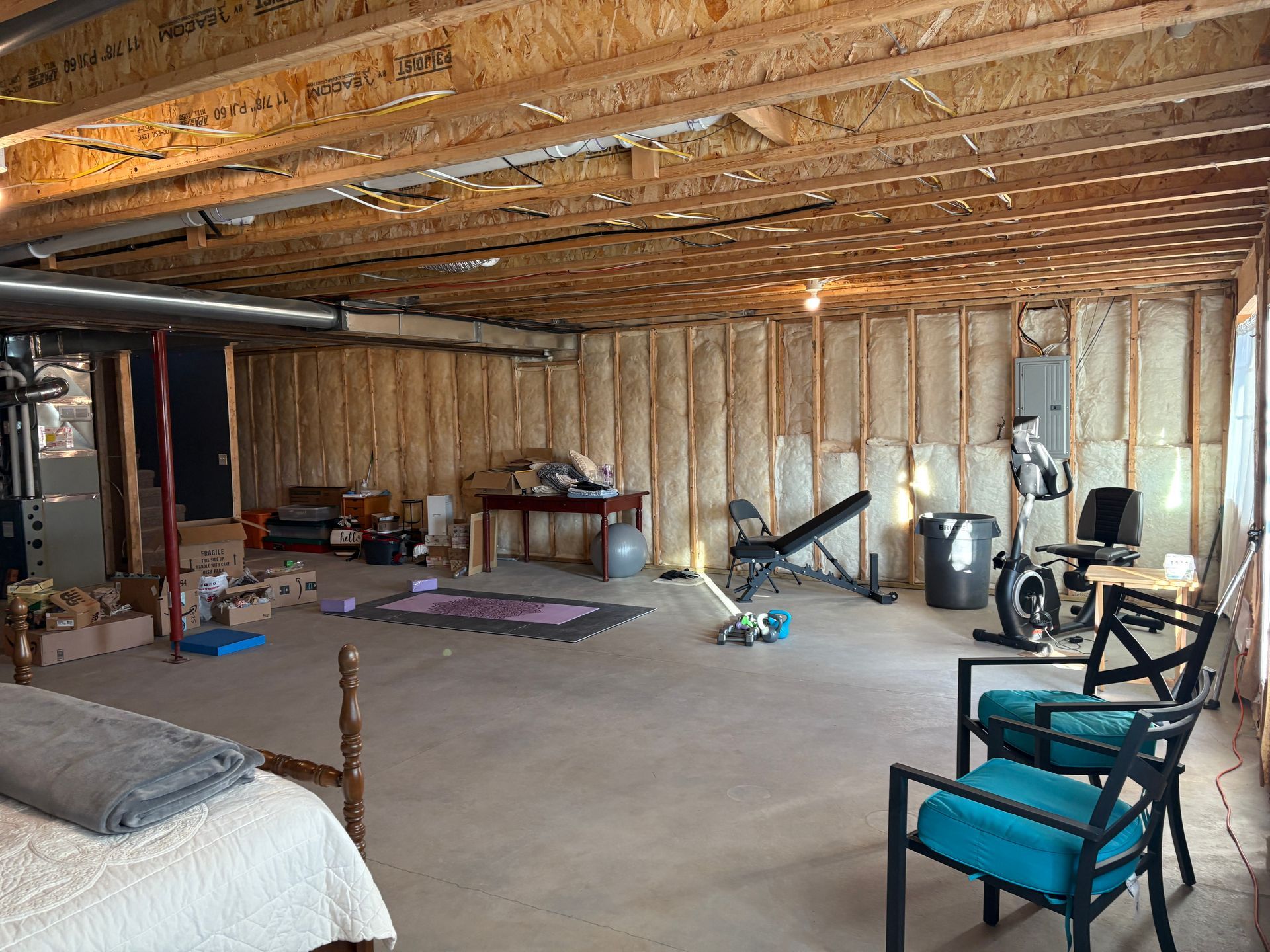 Unfinished basement with exercise equipment, chairs, and a bed. Exposed wooden beams and insulation.