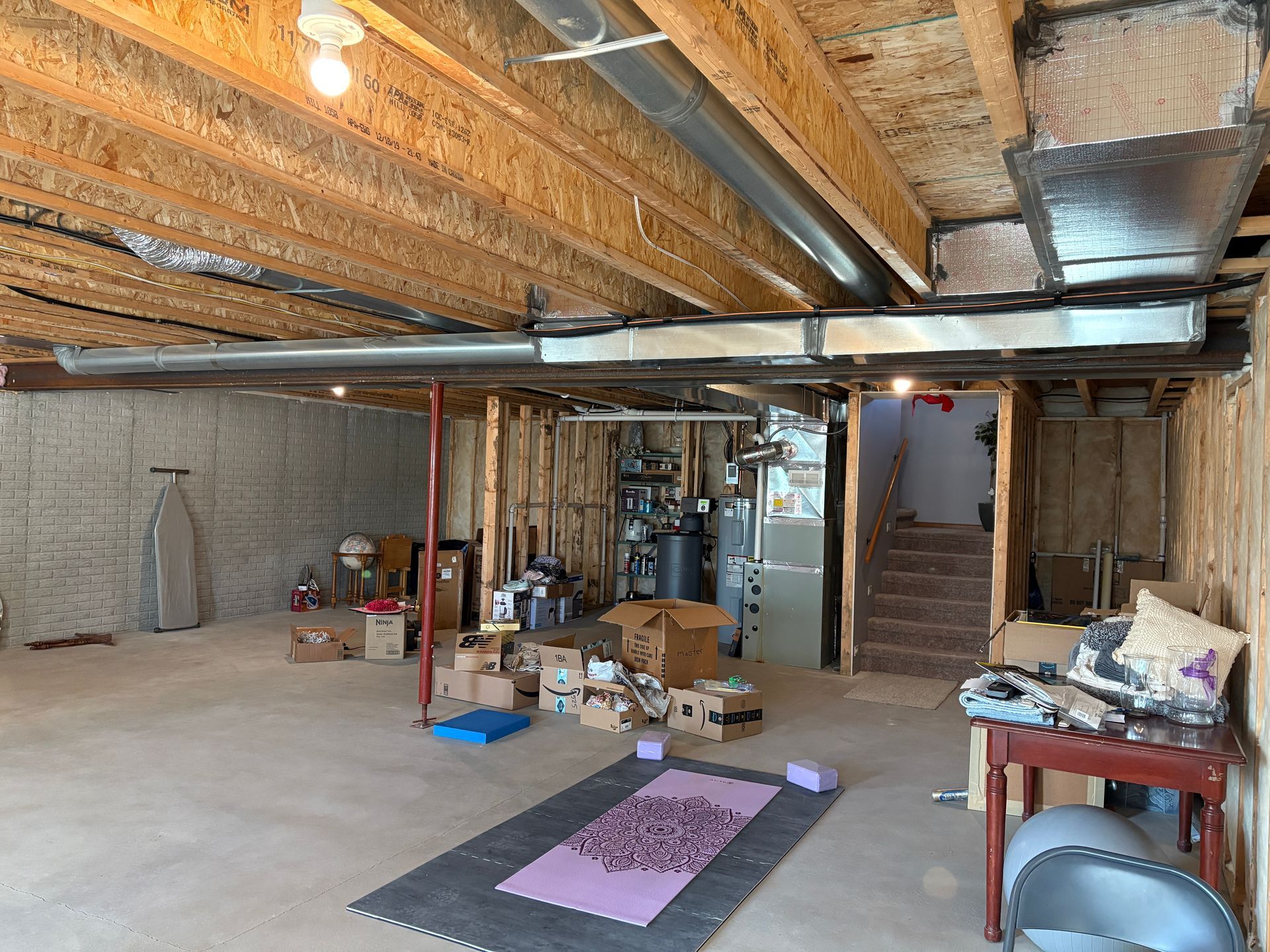 Unfinished basement with exposed beams, ductwork, boxes, exercise mat, and stairs.