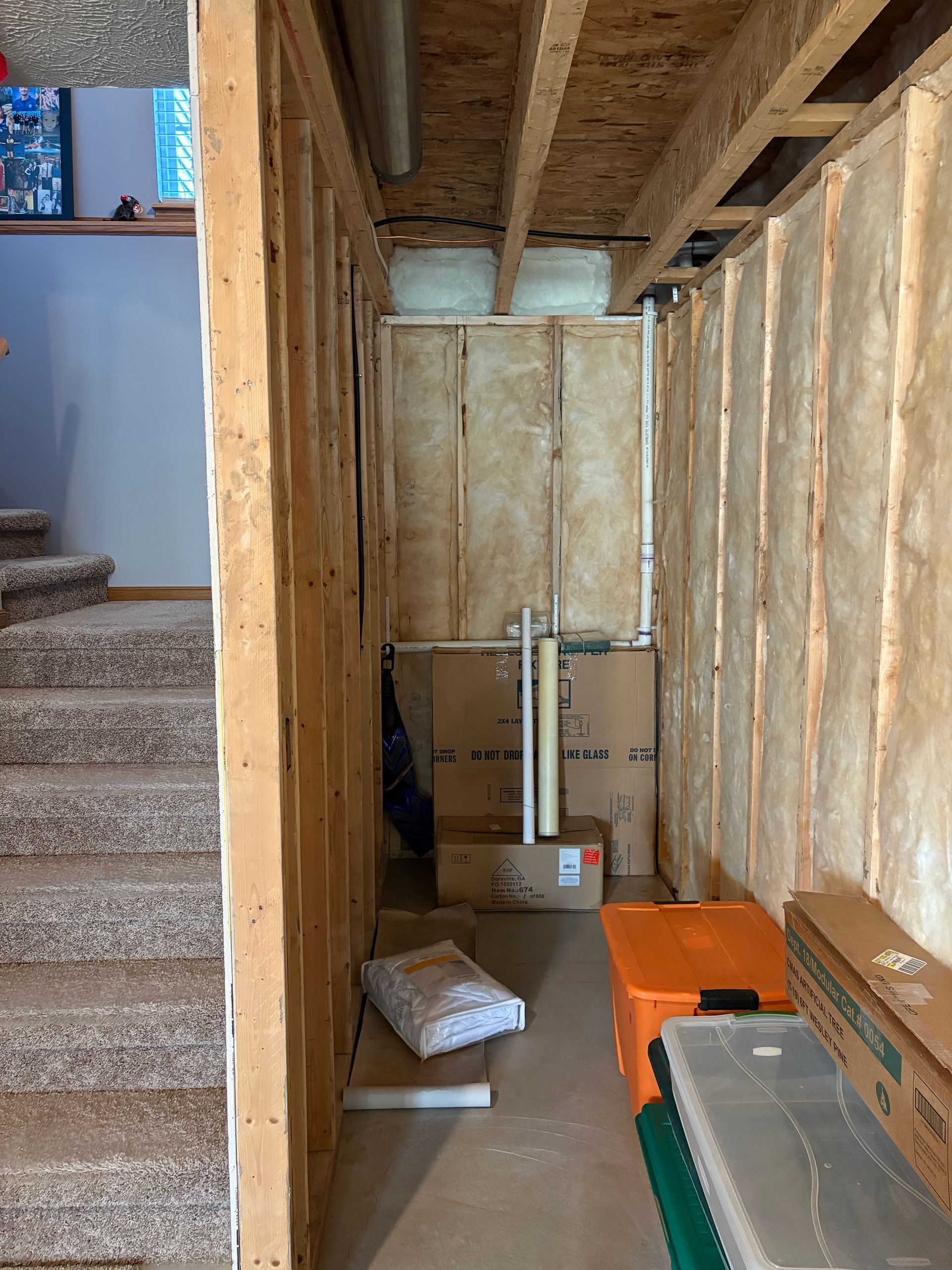 Narrow storage space under stairs with unfinished walls, boxes, pipes, and stairs on the left.