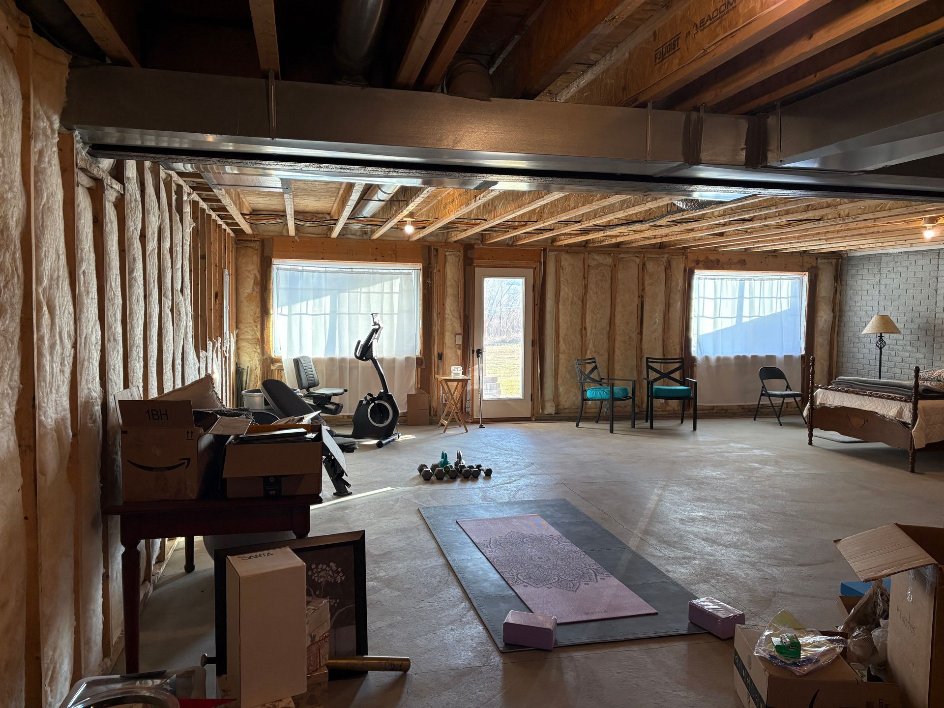 Unfinished basement with exposed beams and insulation; workout equipment, door, and windows; some furniture.