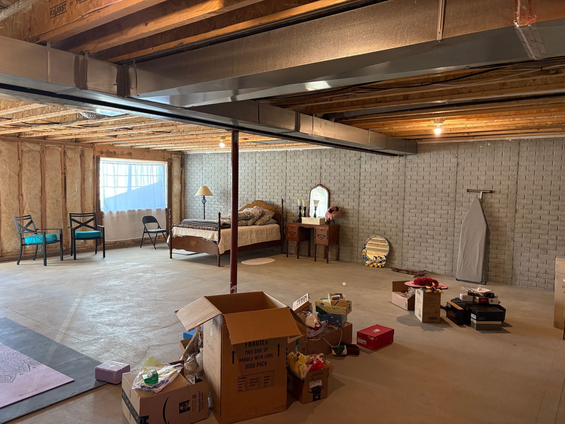 Unfinished basement with a bed, furniture, and storage boxes. Concrete floor, exposed ceiling beams.