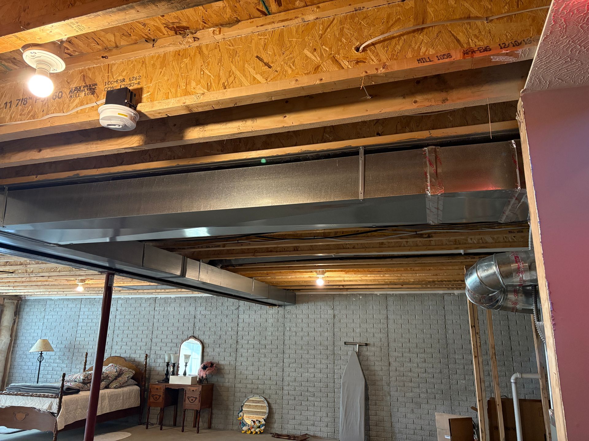 Basement ceiling with ductwork, lights, and exposed wood beams. Furnishings along the gray concrete wall.