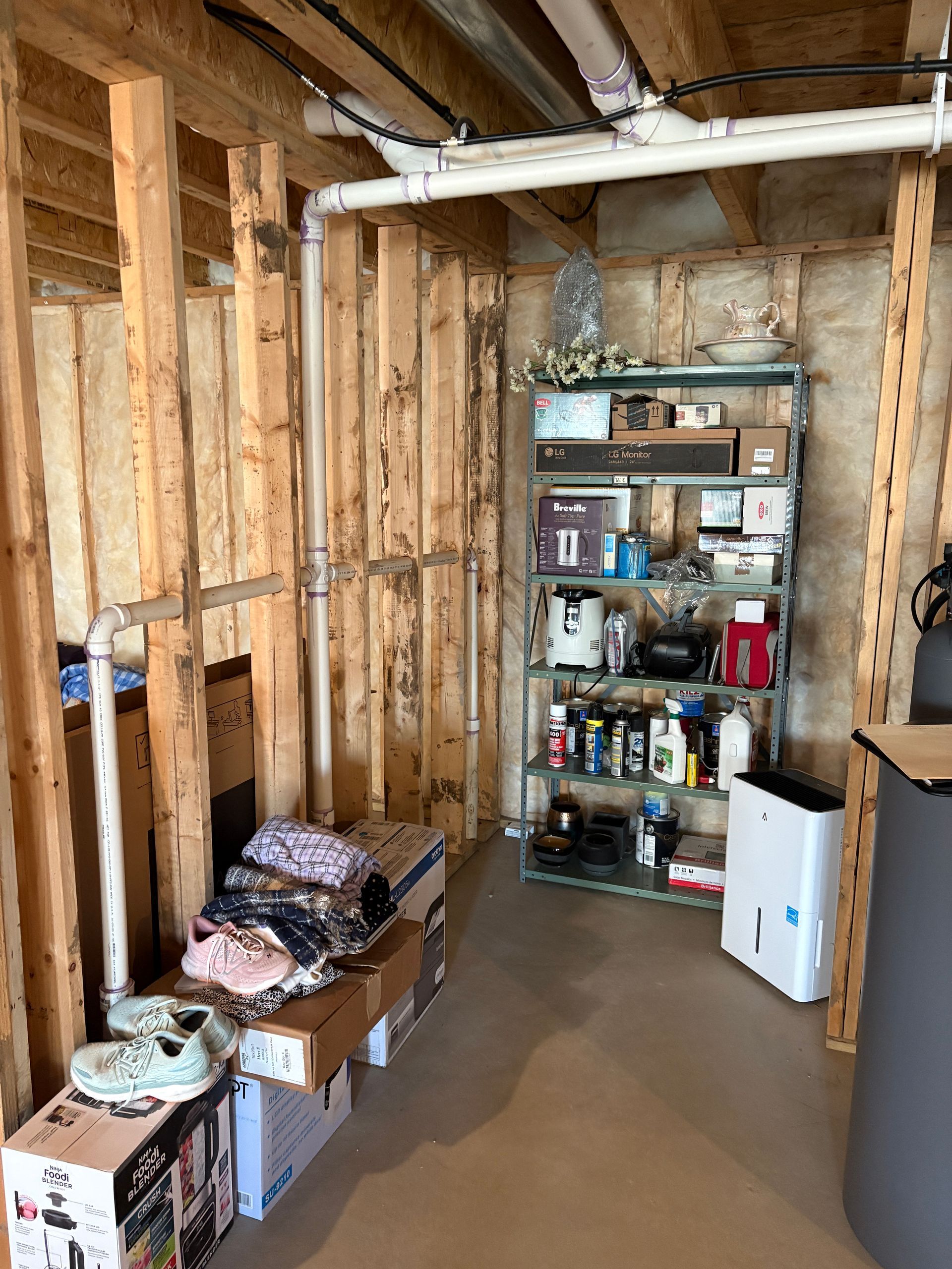 Unfinished basement with exposed wooden studs, shelving unit, clothes, and a dehumidifier.