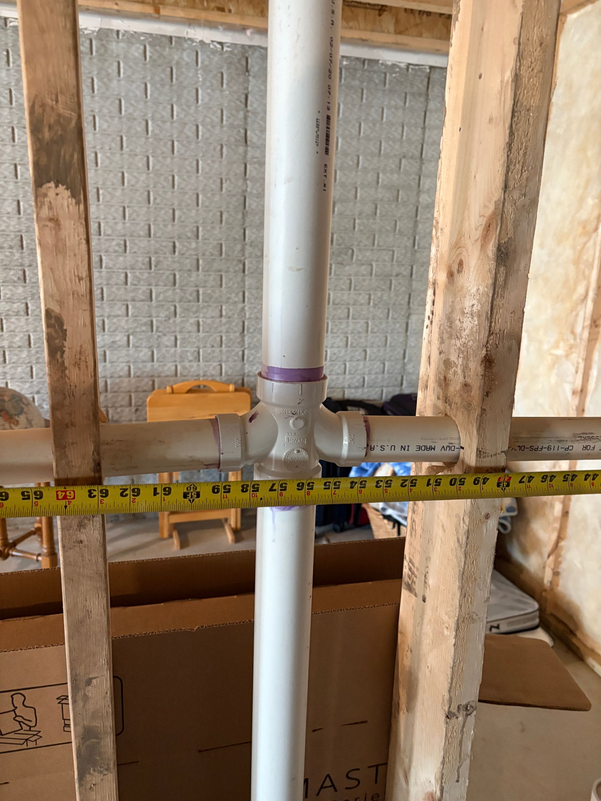 PVC pipes joined with purple fitting between wooden framing. A measuring tape lies across pipes.
