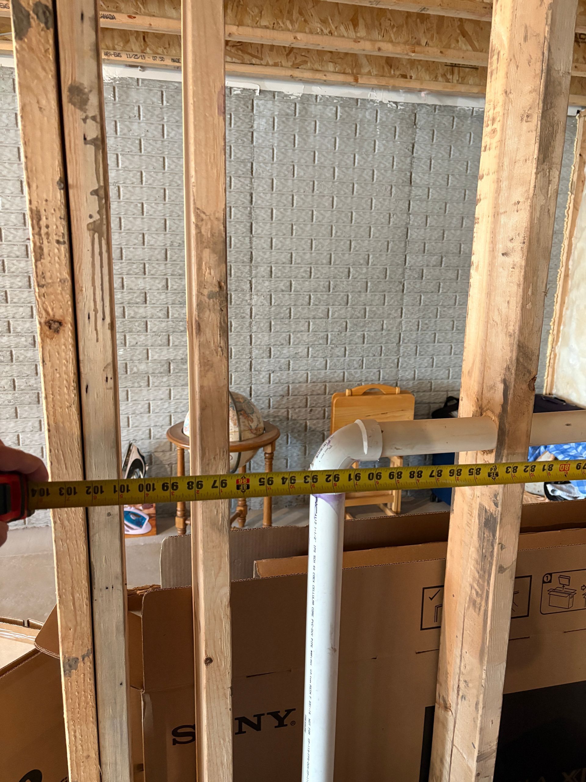 A PVC pipe in a wood-framed wall, with a measuring tape indicating a length in an unfinished space.