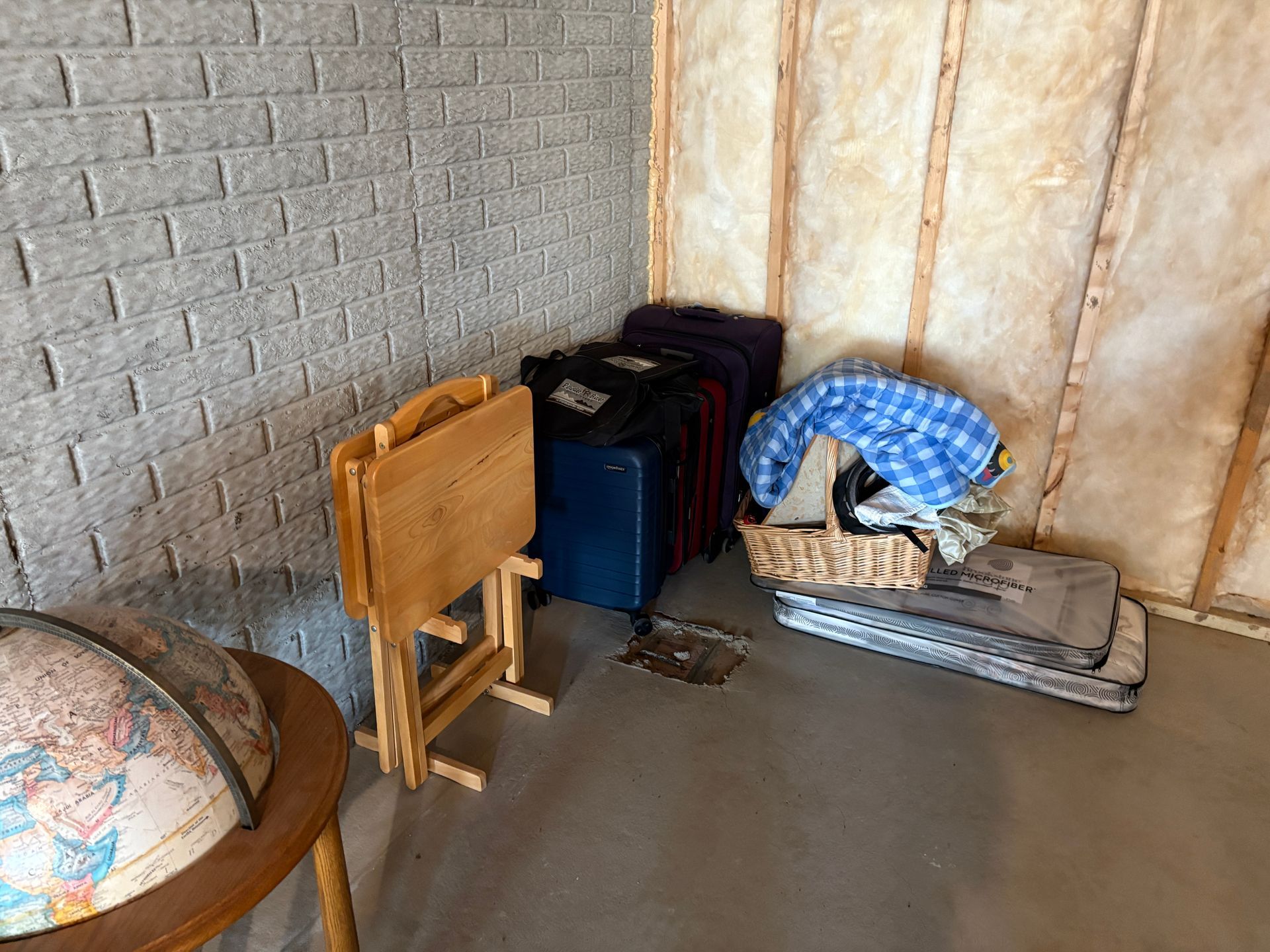 A corner with luggage, table, globe, and a basket of items against a gray brick wall and light wood frame.