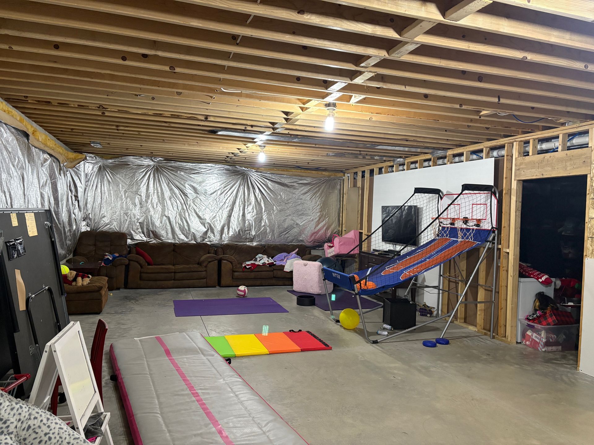 Basement room with couch, gym equipment, and exposed ceiling. Silver insulation on one wall.