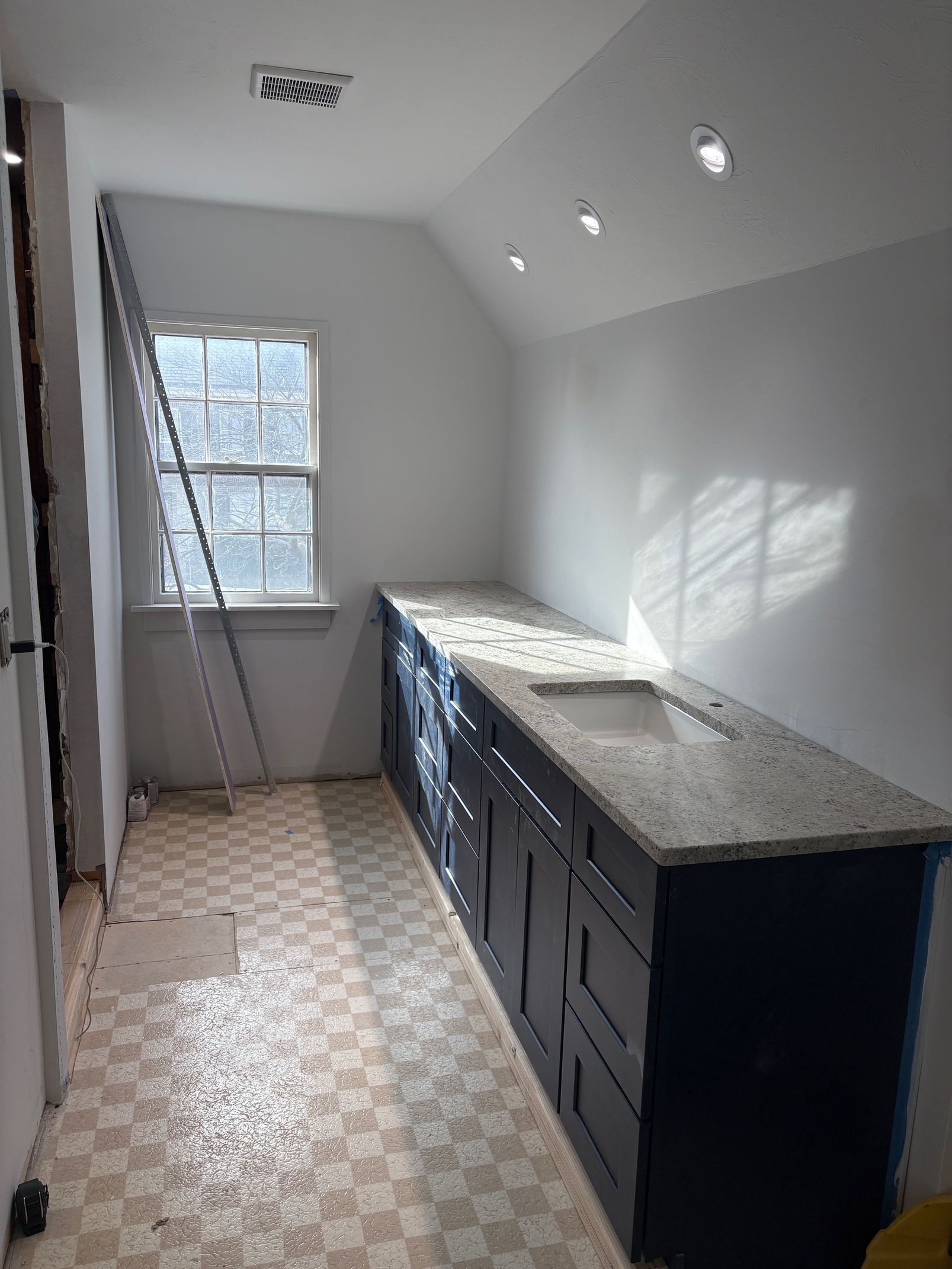 Renovated kitchen with blue cabinets, granite countertop, window, and ladder. Walls are white; flooring is patterned.