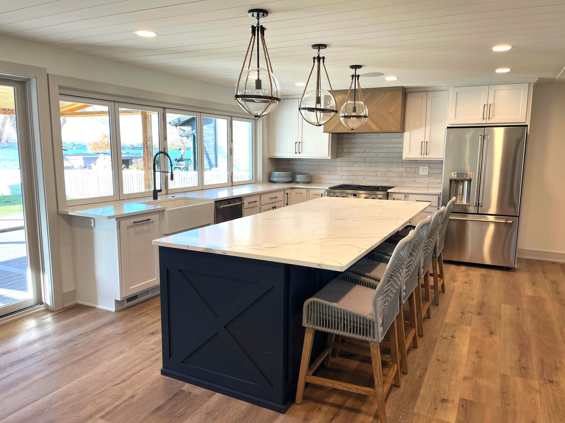 A modern kitchen with a navy island, white countertops, and wood flooring. Large windows and pendant lights are visible.
