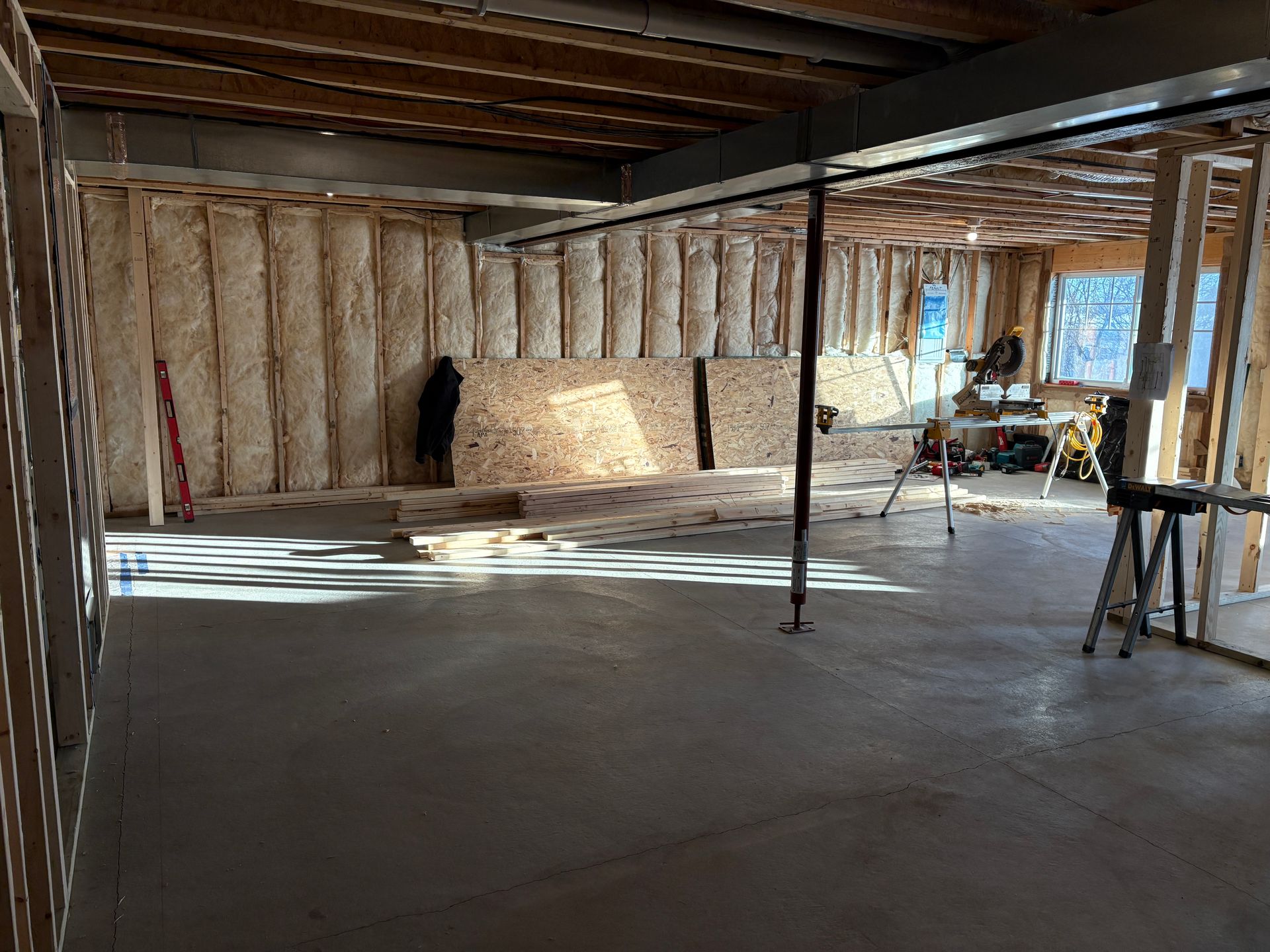 Basement under construction with concrete floor, exposed studs, insulation, and tools. Natural light streams in.