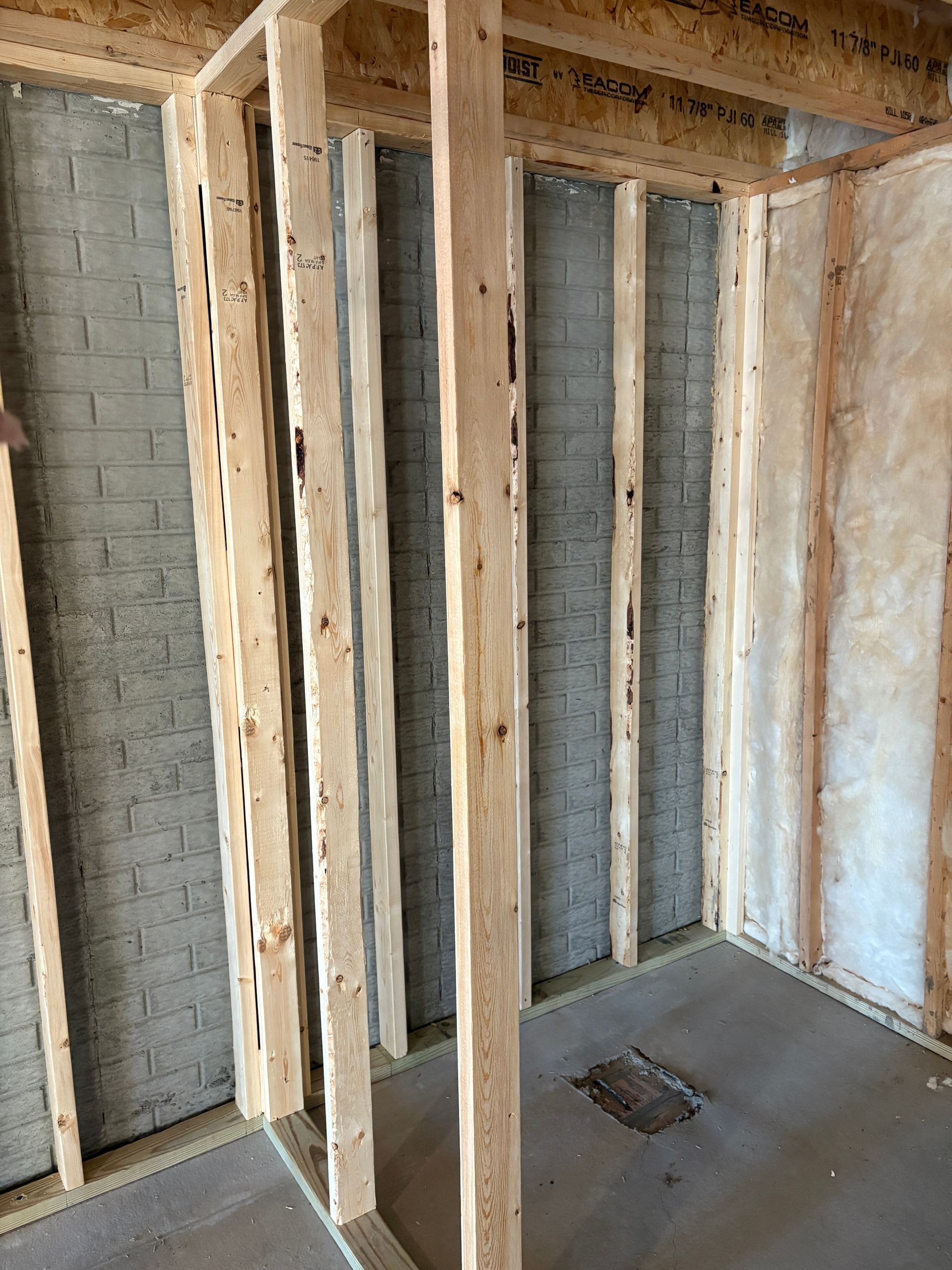 Framing of a room. Studs of wood against a brick wall and insulated wall. Floor is concrete.