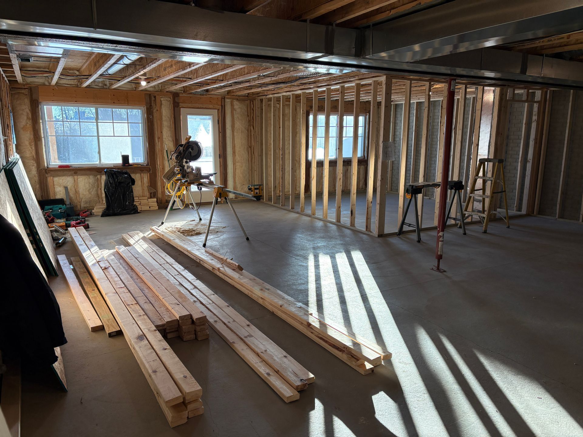 Basement renovation in progress, interior framing, lumber, tools, window, door, and concrete floor.