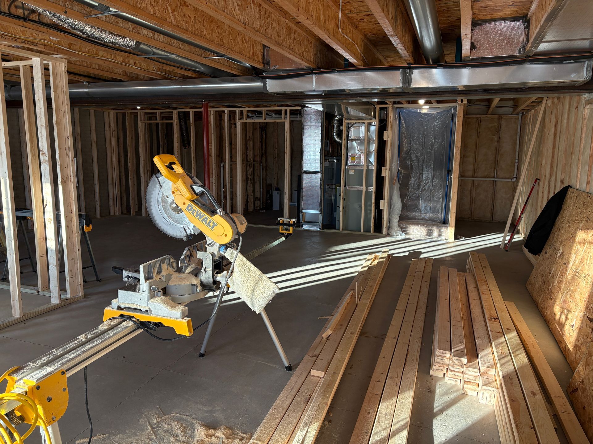 A miter saw with lumber in a basement under construction; wooden framing and ductwork are visible.