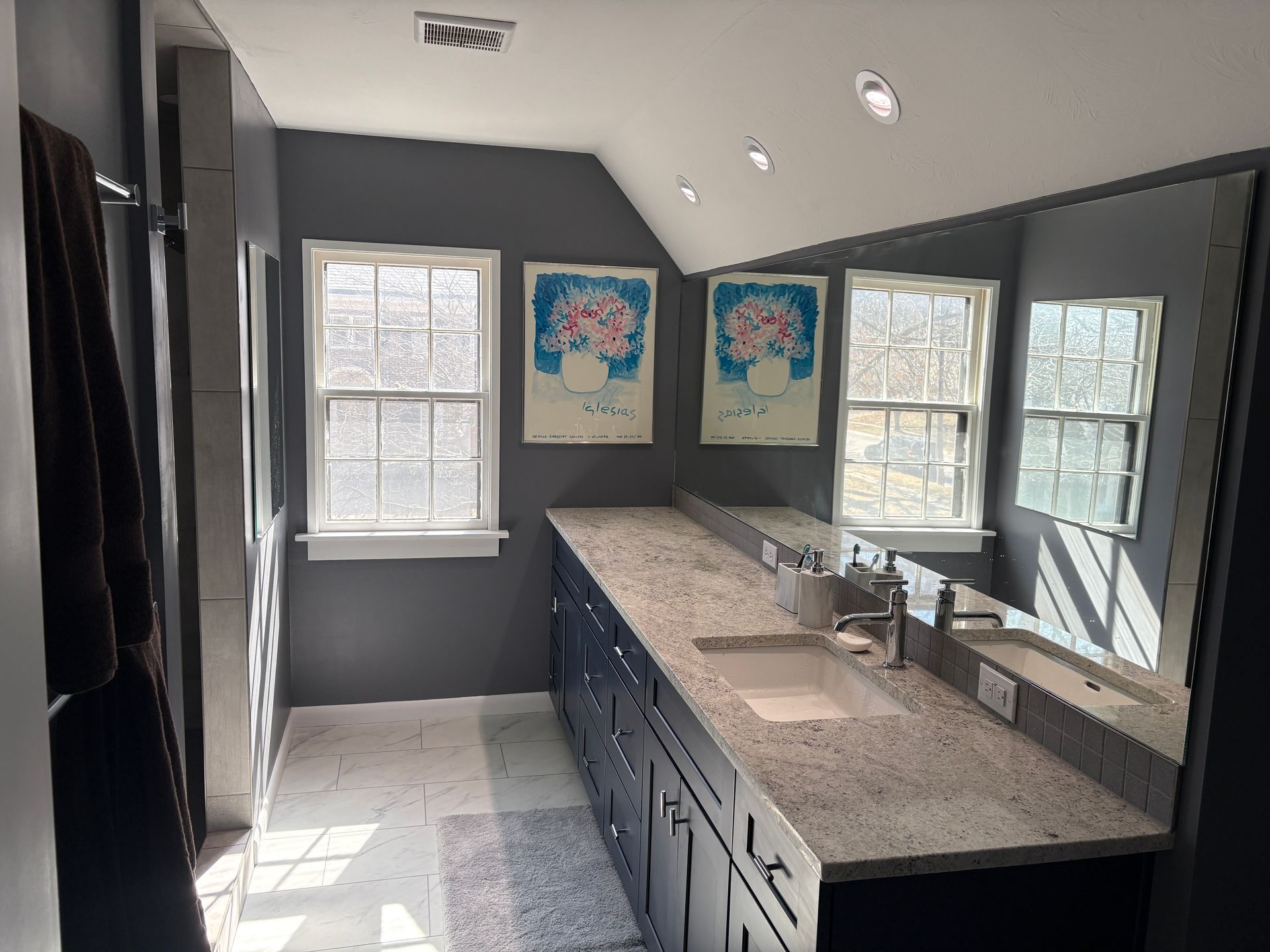 Dark gray bathroom with long vanity, two windows, art prints, and a large mirror.