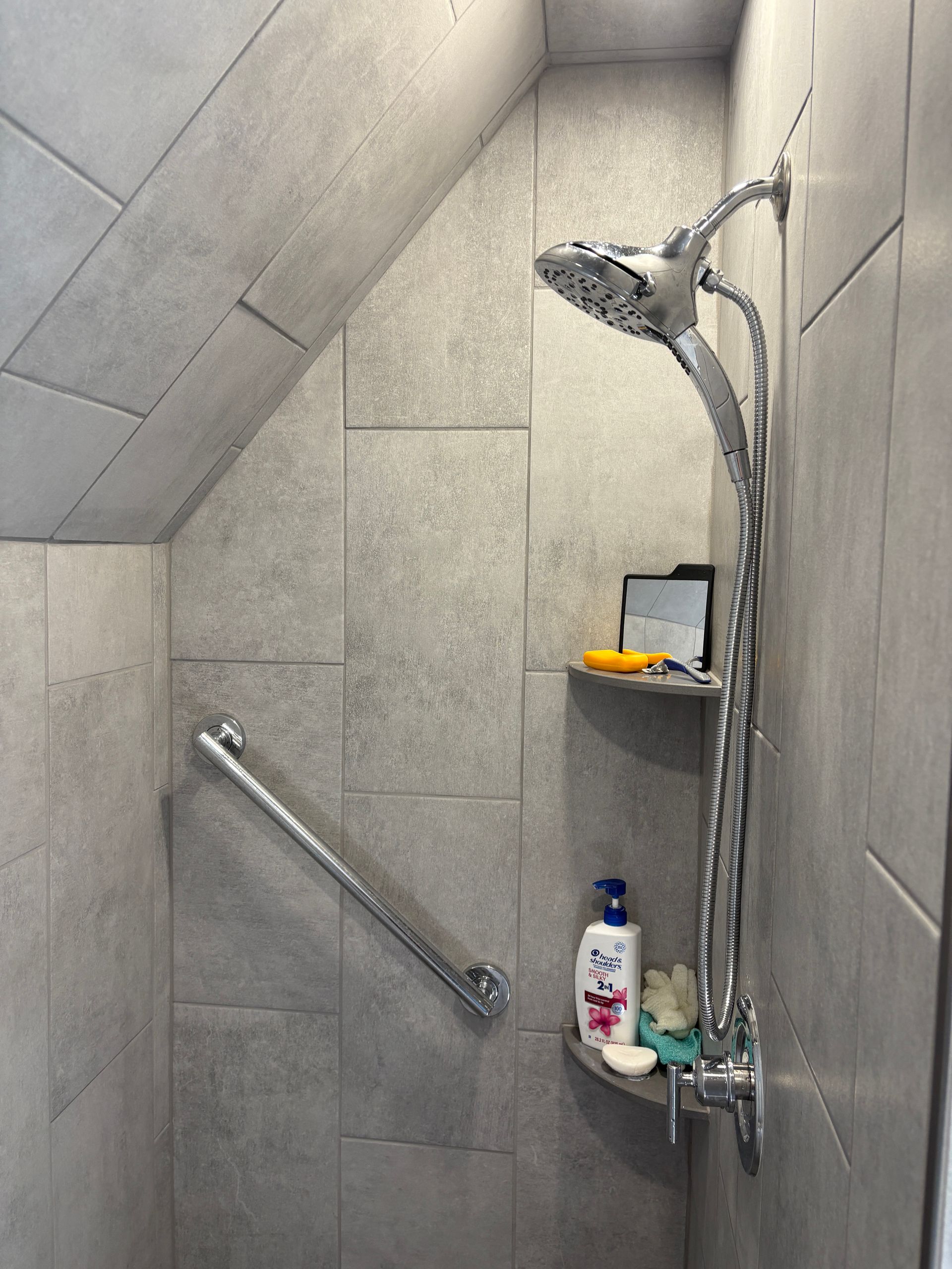 Gray-tiled shower with a handheld showerhead, grab bar, and built-in shelf with toiletries. Angled ceiling.