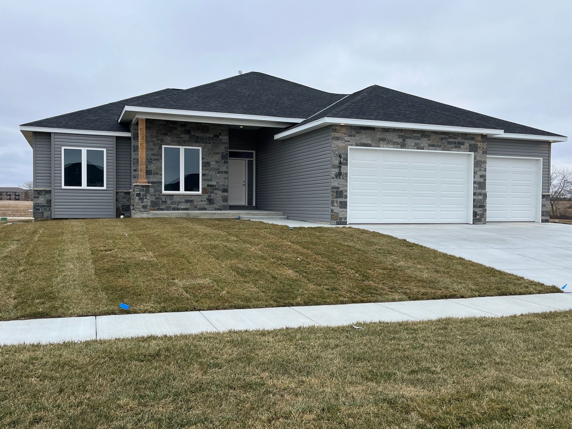 Gray house with stone and siding, two-car garage, and a lawn under an overcast sky.