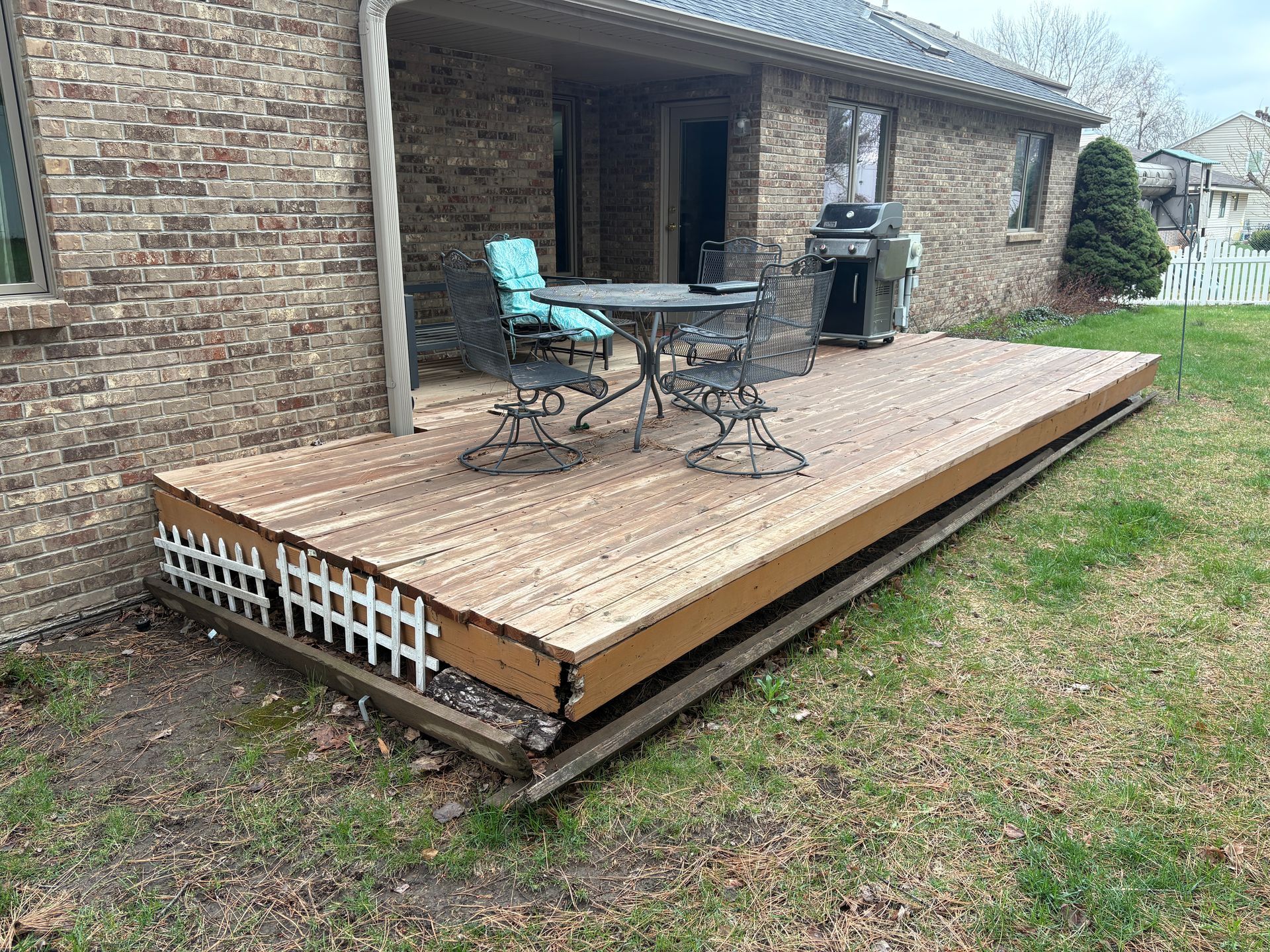 Backyard deck with furniture, grill, and damaged trim against a brick house.
