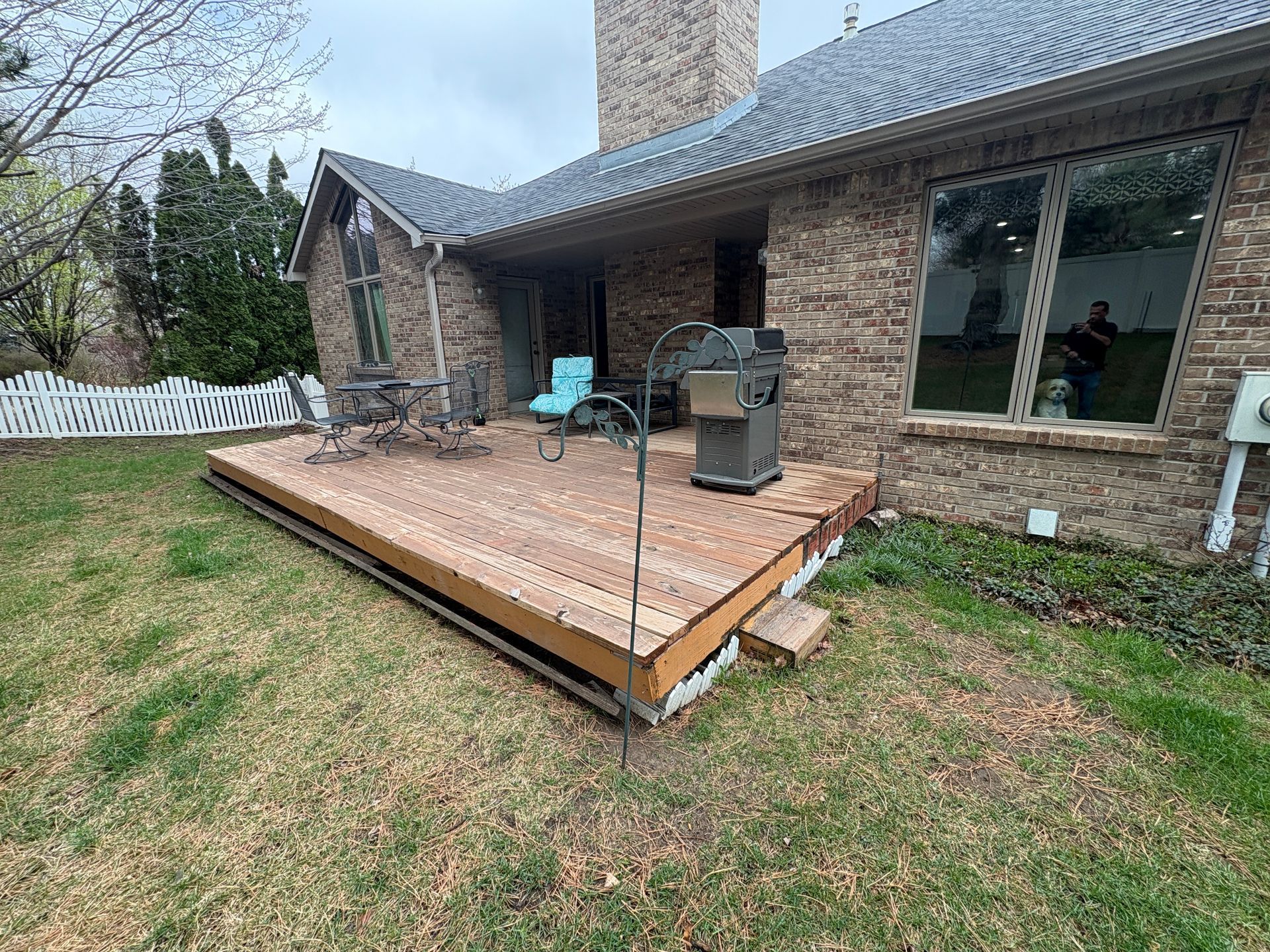 A wooden deck with grill and chairs next to a brick house and lawn.