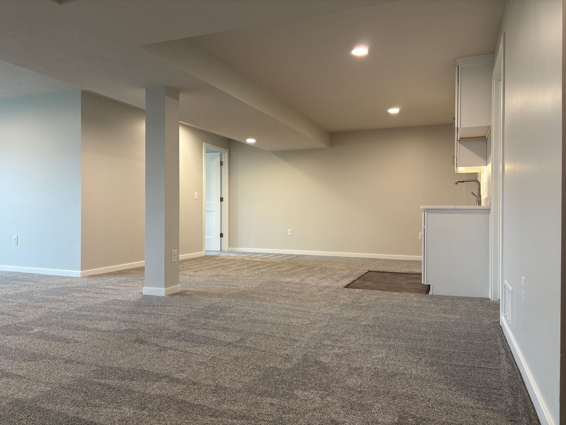 Empty basement with gray carpet, light gray walls, a white cabinet and a support beam.