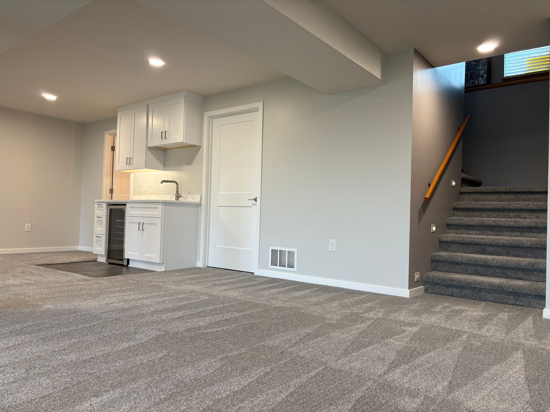 Basement room with a kitchenette, stairs, and carpet in neutral tones.