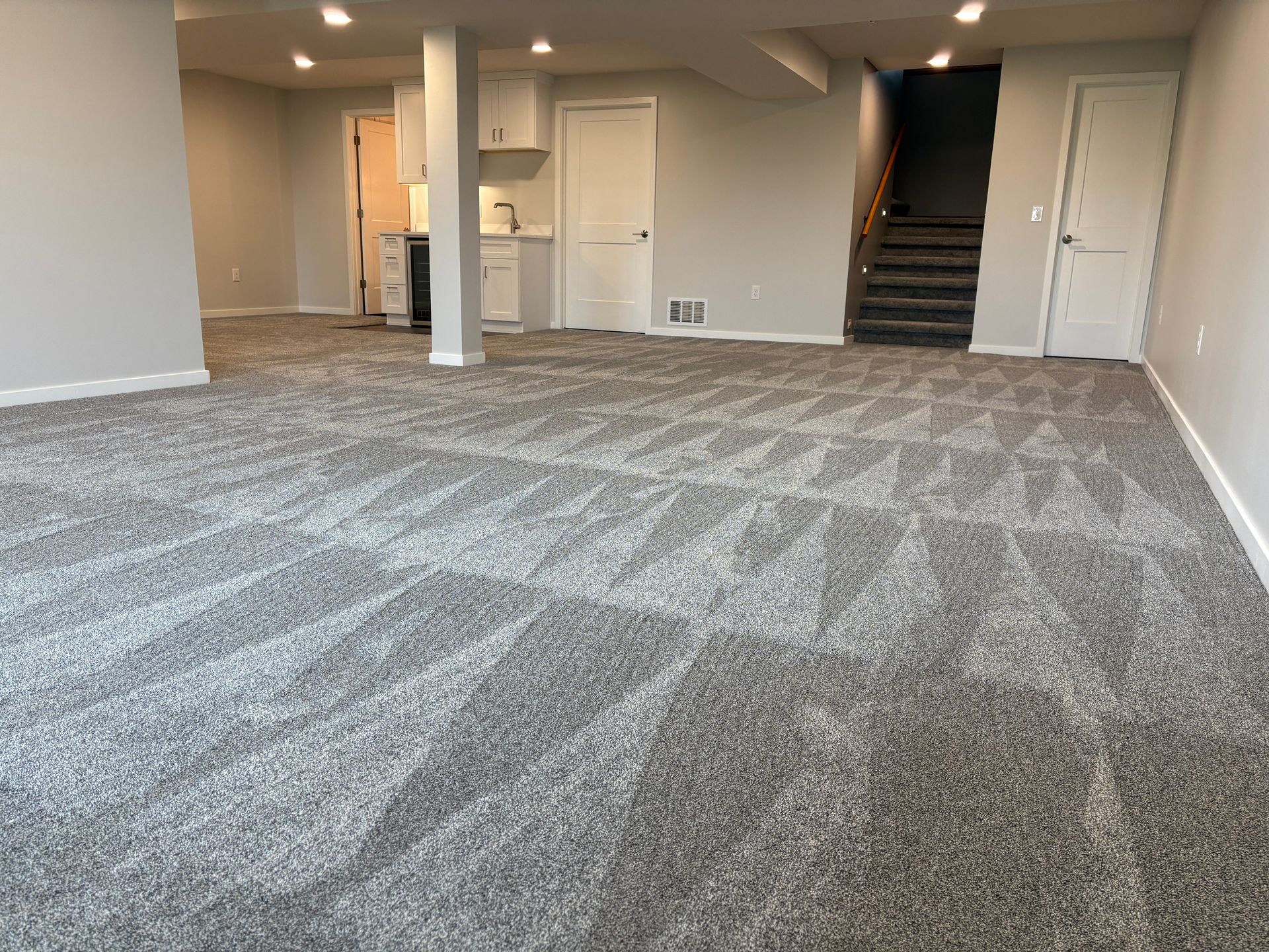 Gray carpeted basement with built-in cabinets and stairs. White walls, recessed lighting, and a door.