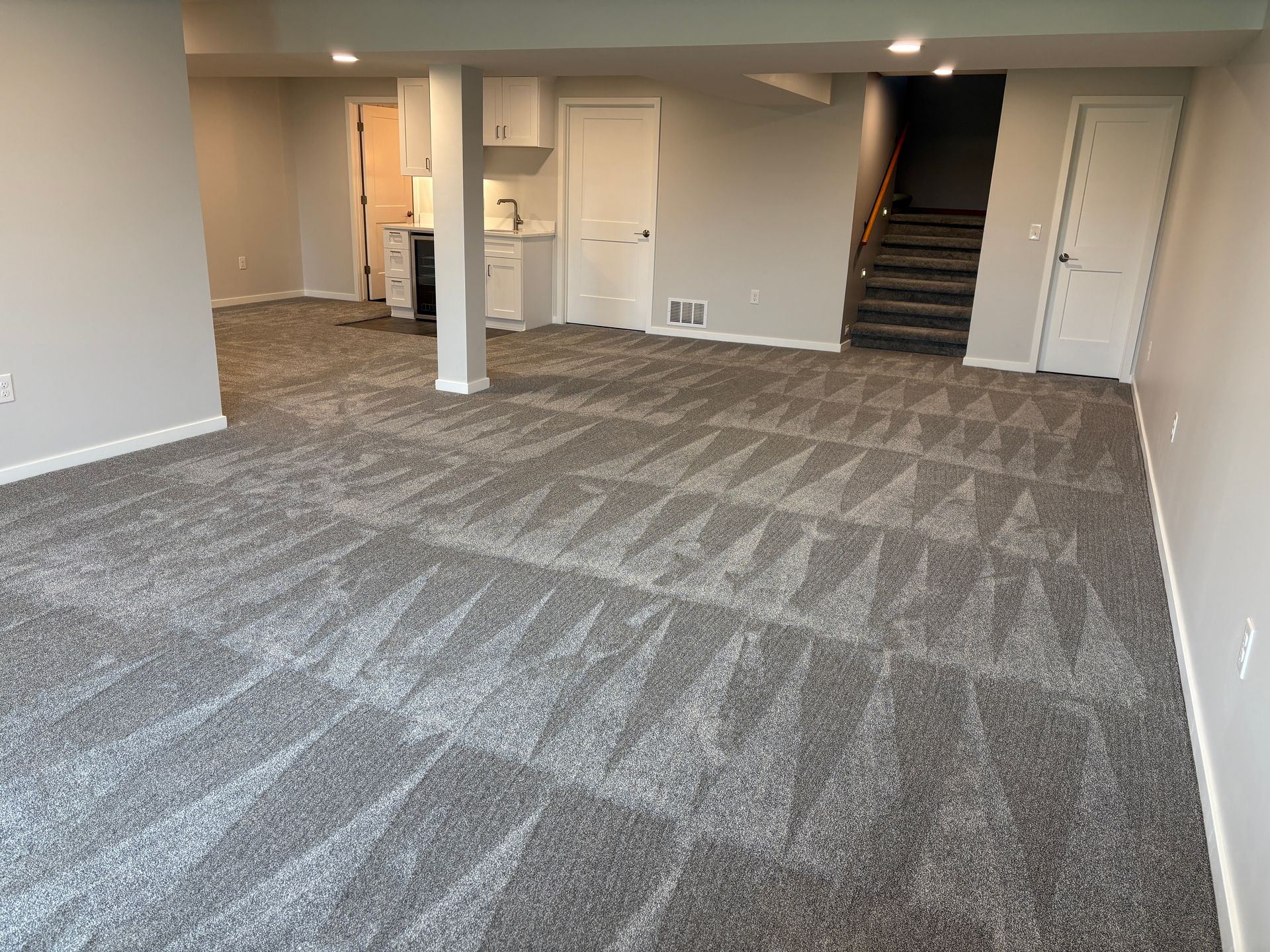 A spacious basement with new grey carpet, neutral walls, and a small kitchenette near a staircase.